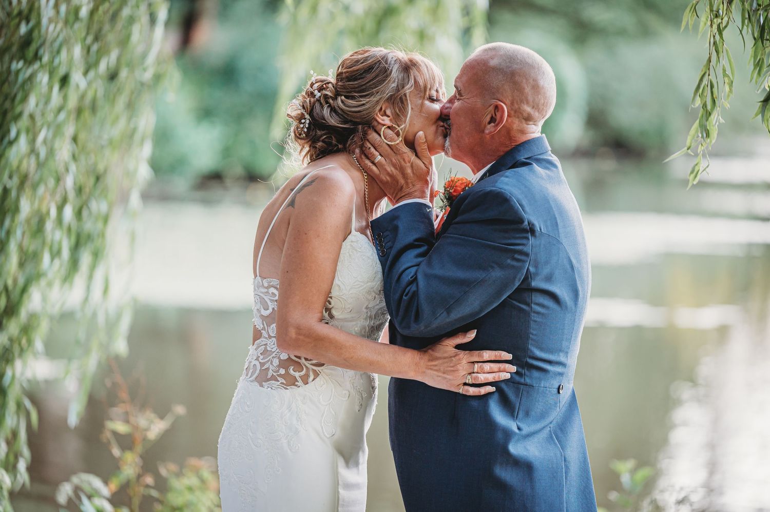 A romantic wedding couple shares a loving kiss under graceful willow branches beside a tranquil pond.