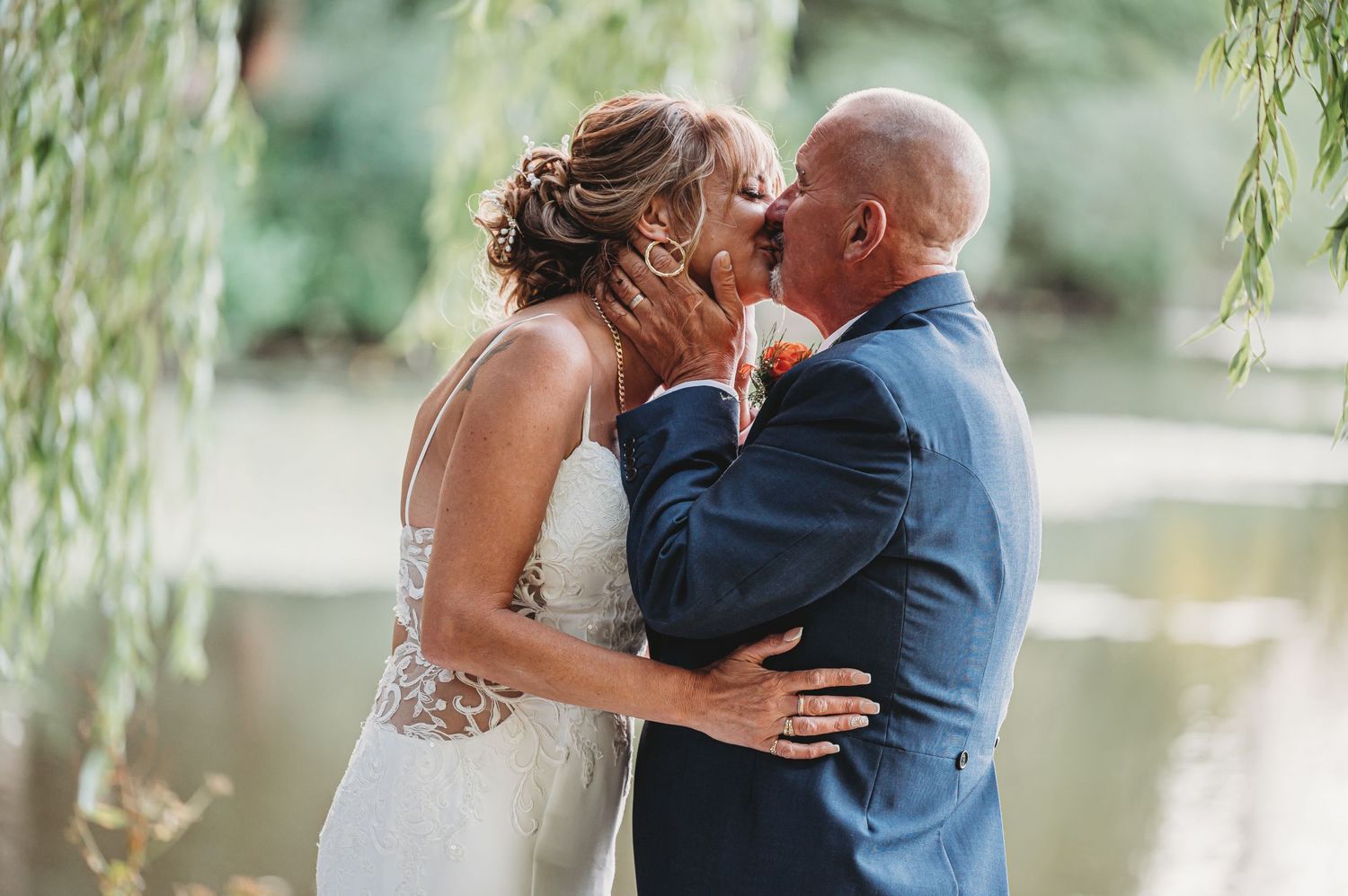 A romantic wedding kiss under drooping willow branches near a pond on a summer day.