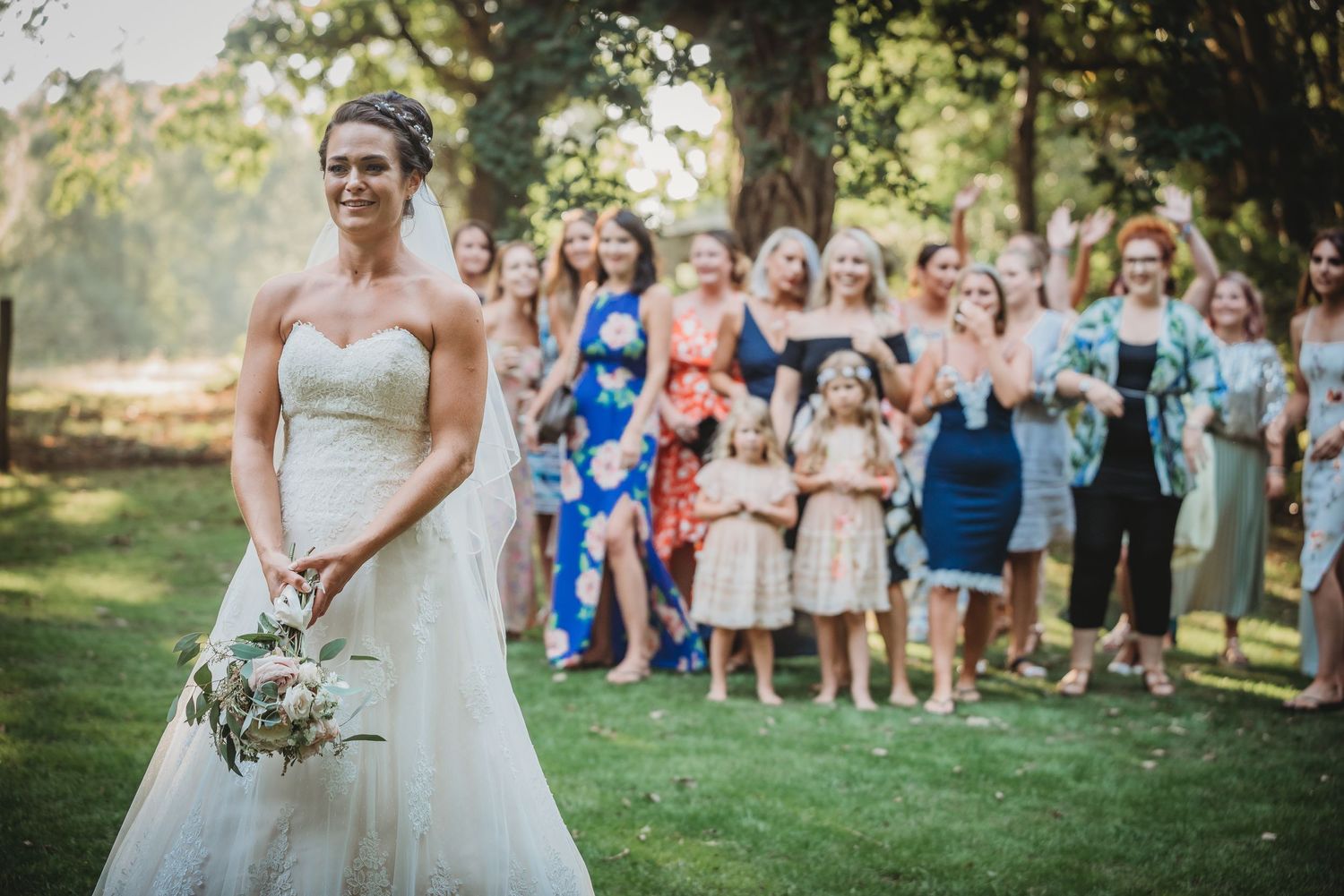 A bride in a white gown holds a bouquet while wedding guests stand in a line on the lawn behind her.