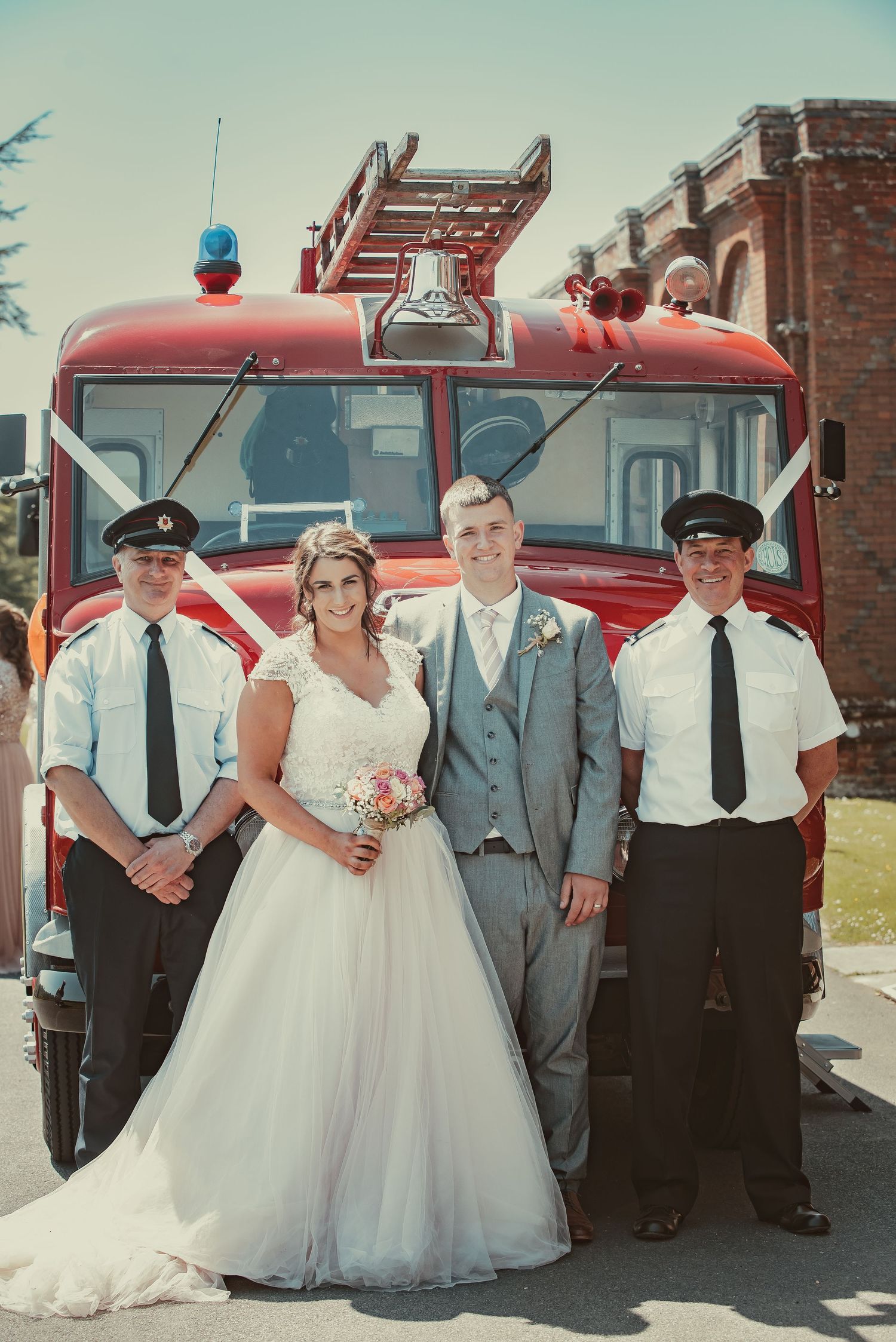 A vintage red fire truck serves as a unique backdrop for a wedding photo on a sunny day.