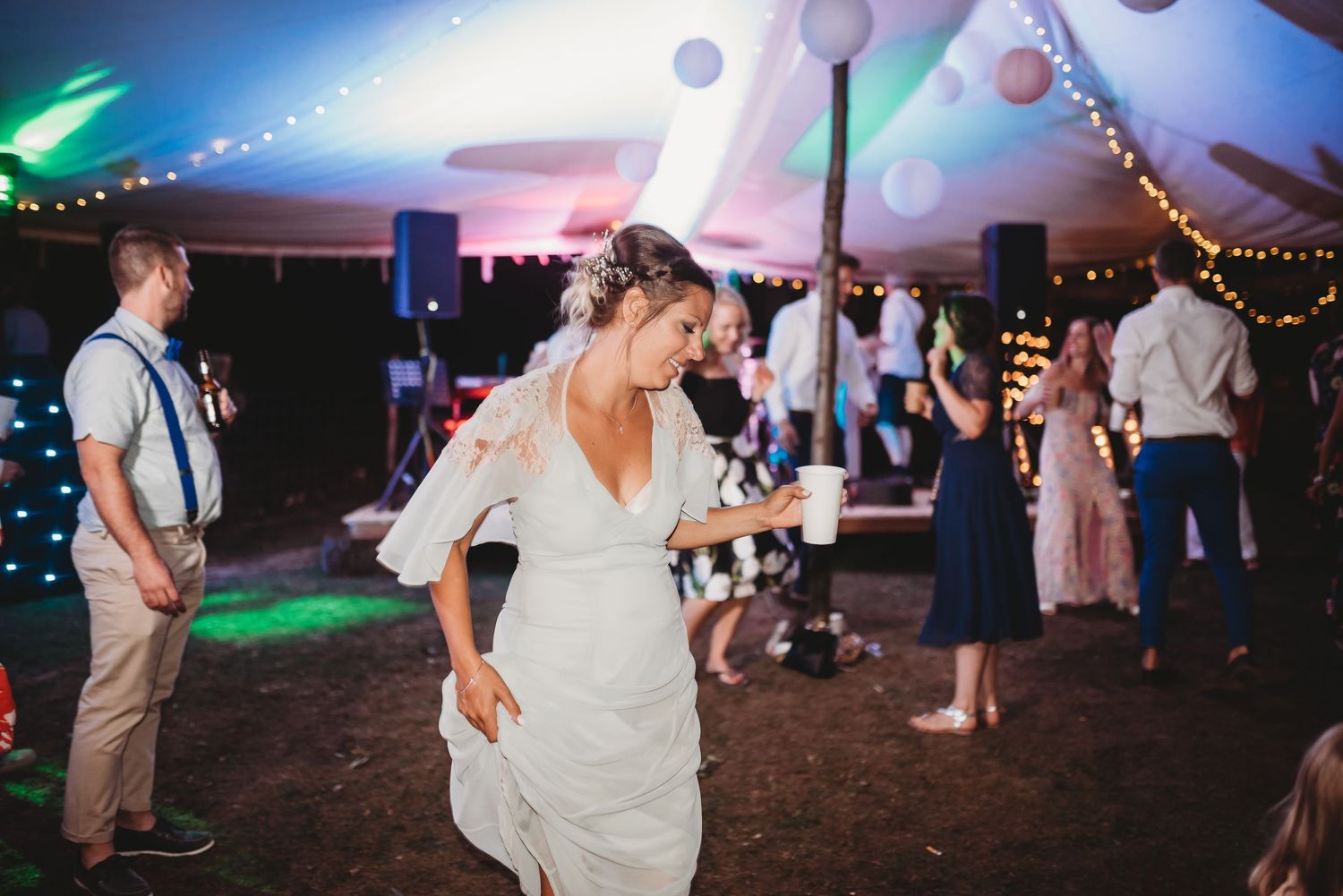Wedding guests dance and celebrate under a tent decorated with string lights and hanging decorations at an evening reception.