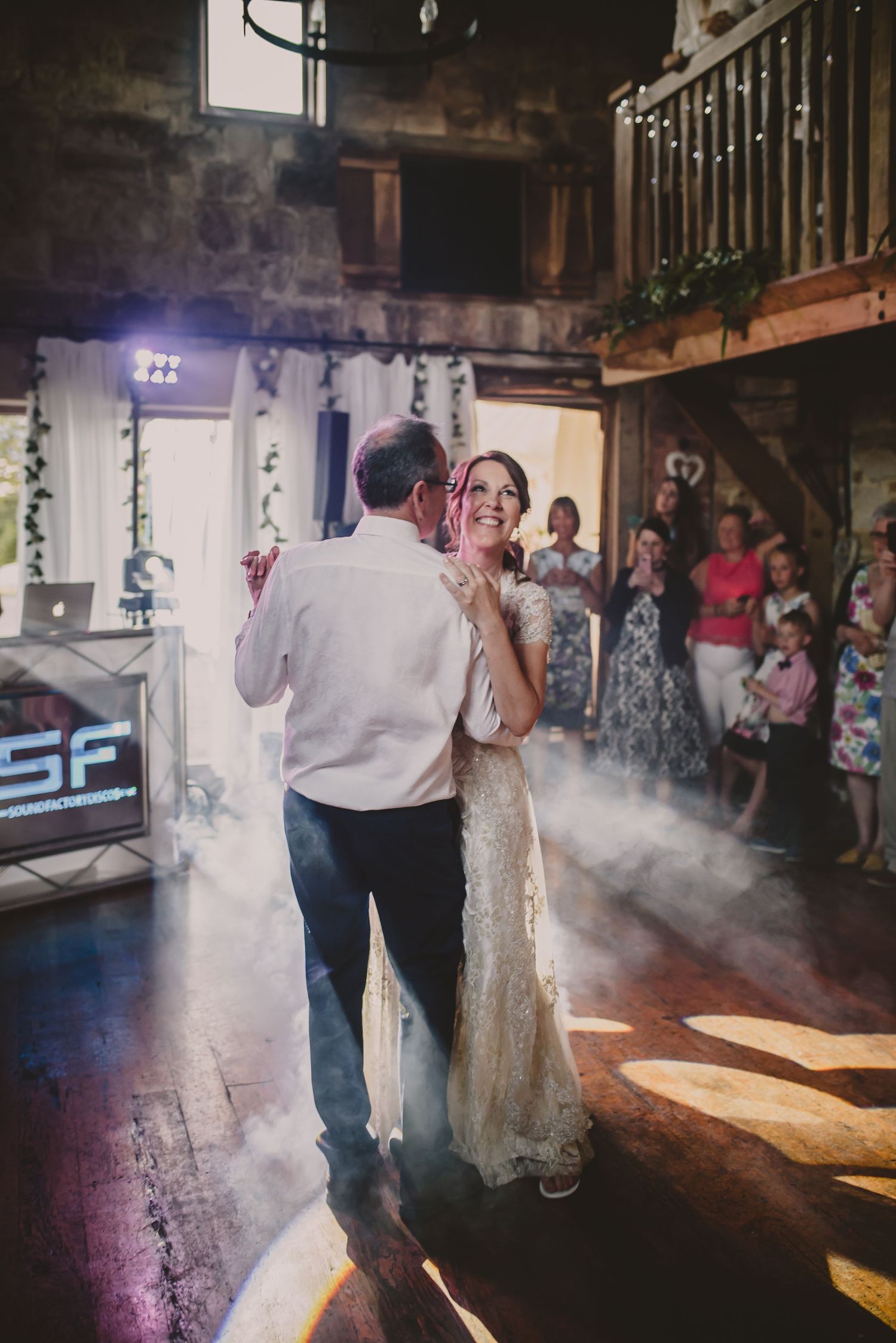 A couple shares their first dance as newlyweds on a misty dance floor with guests watching from the background.