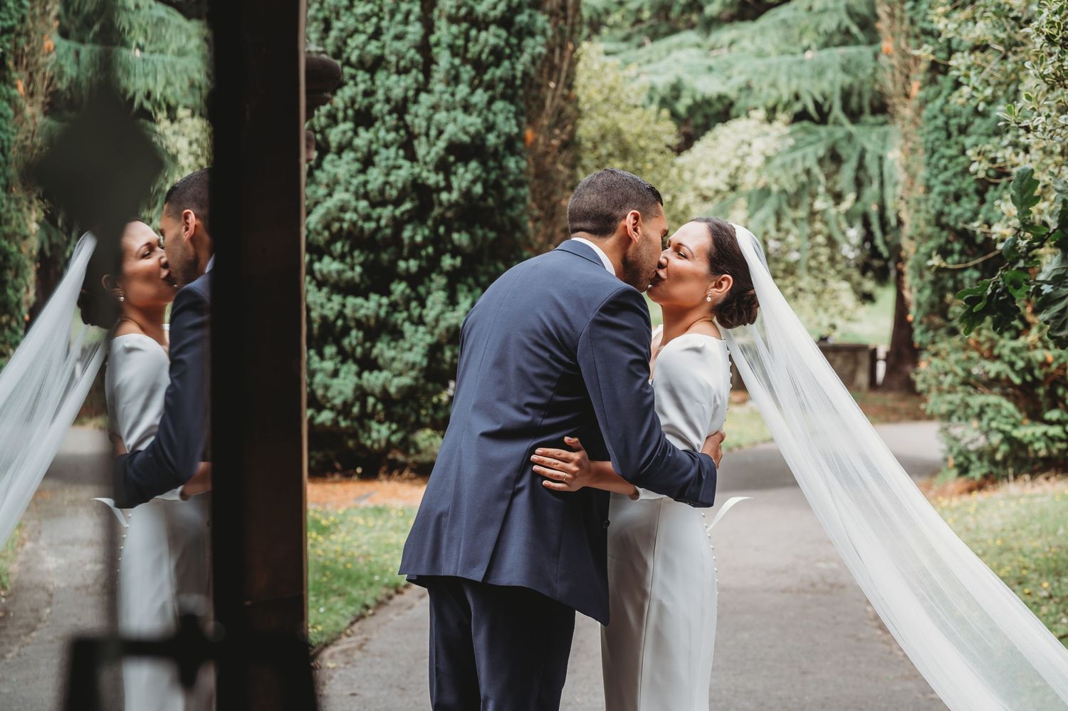 A couple shares a romantic kiss on their wedding day in a lush garden setting surrounded by greenery.