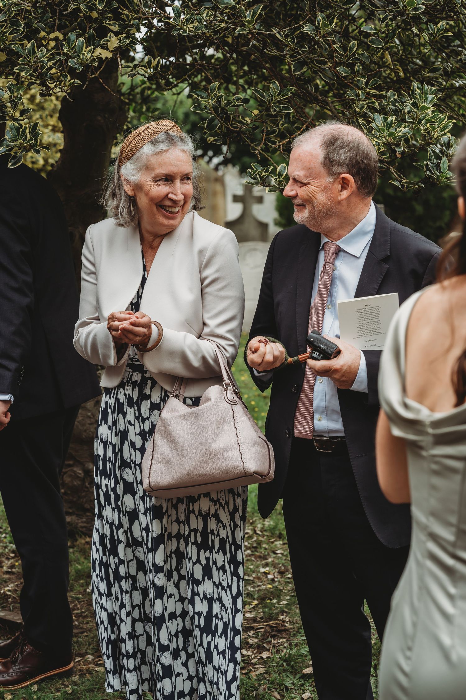 Well-dressed guests sharing a laugh at an outdoor social gathering under trees.