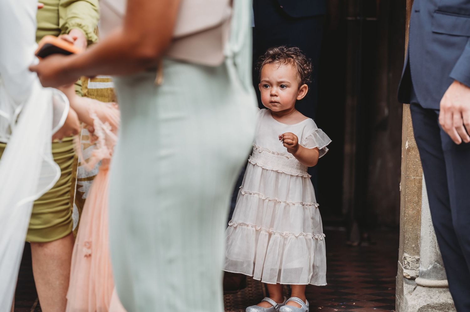 A small child in a ruffled cream dress stands curiously among adults at an indoor gathering.