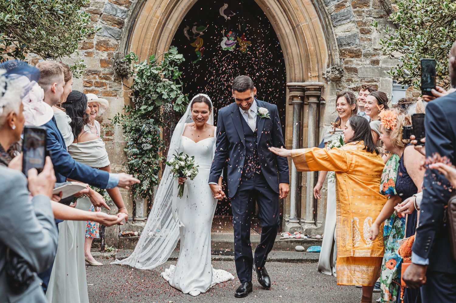 Happy newlyweds exit stone church as wedding guests shower them with confetti on beautiful countryside ceremony.