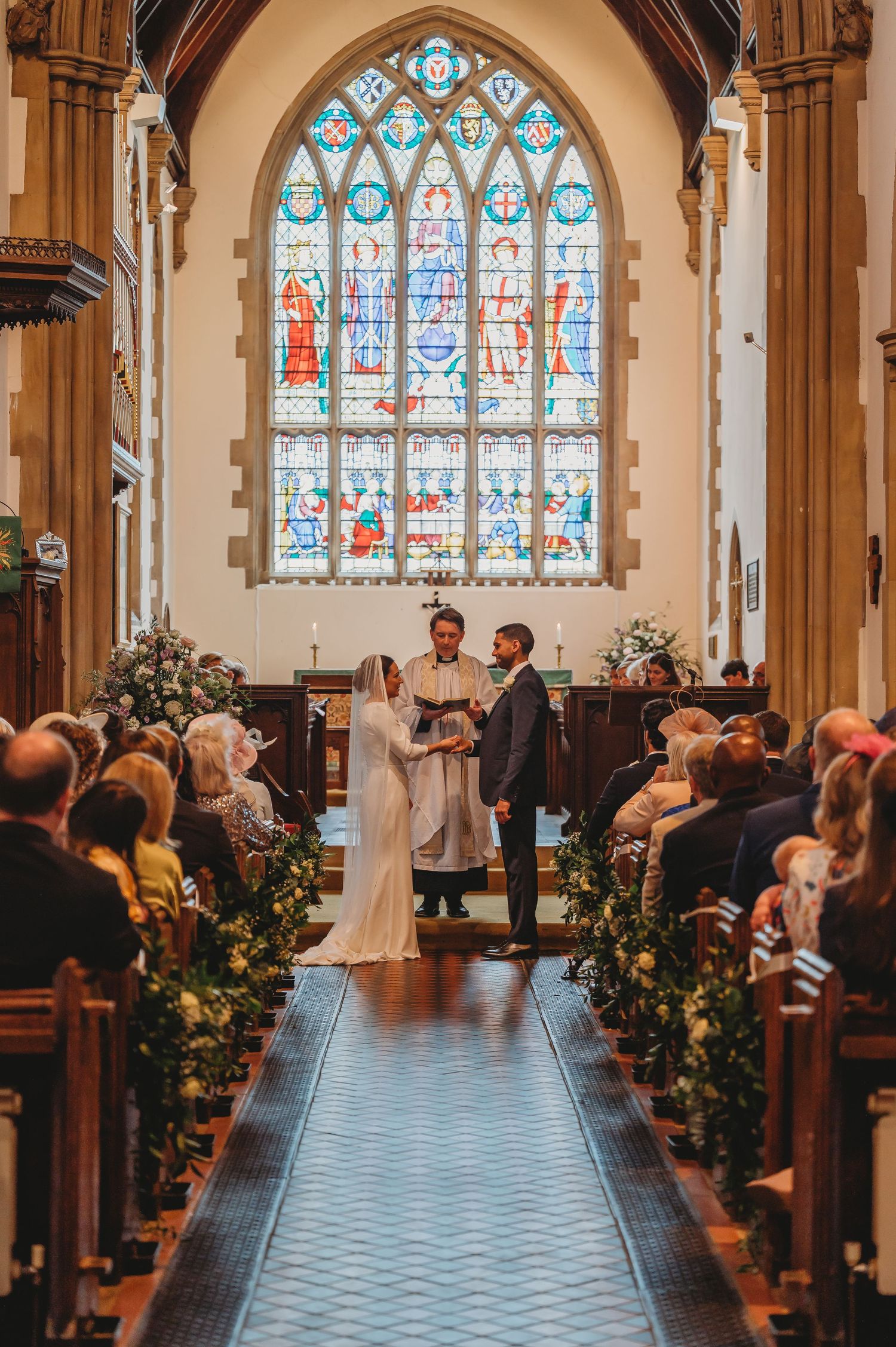A wedding ceremony takes place in a beautiful church with stained glass windows and floral decorations along the aisle.