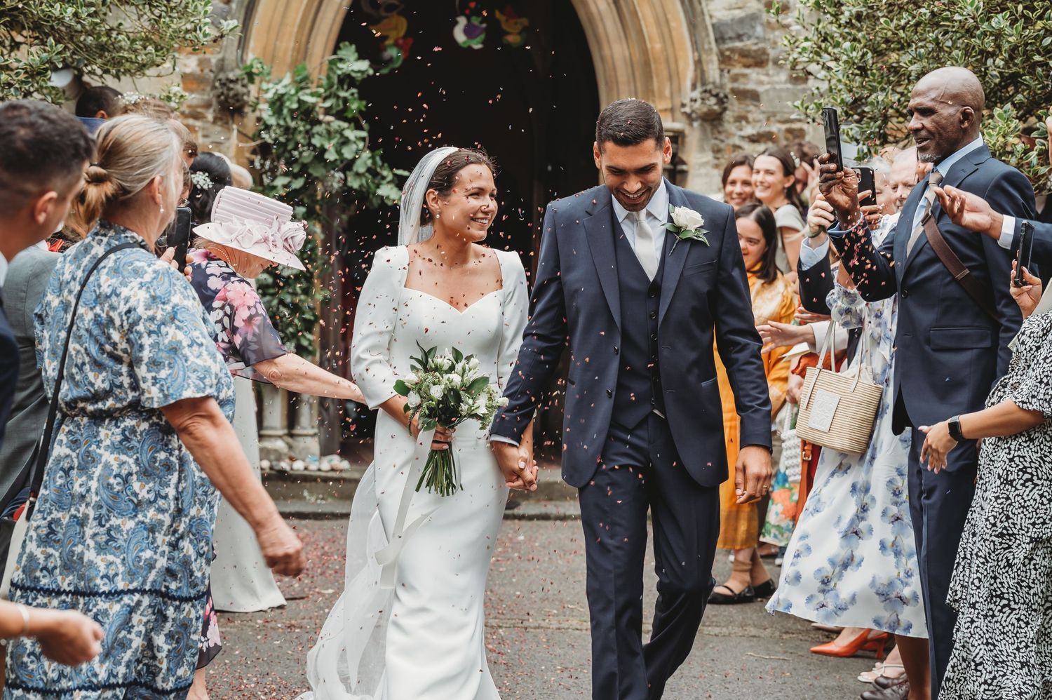 Newlyweds exit church after wedding ceremony while guests throw confetti in celebration on stone archway steps.