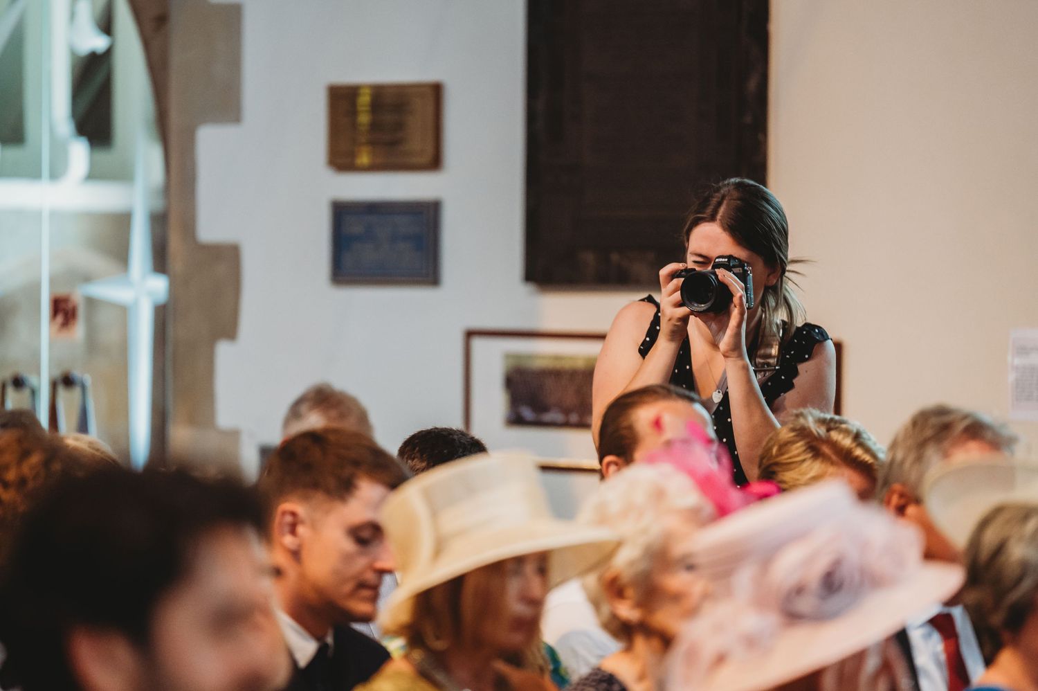 A photographer captures a crowded event with attendees wearing fancy hats and elegant attire in an indoor venue.