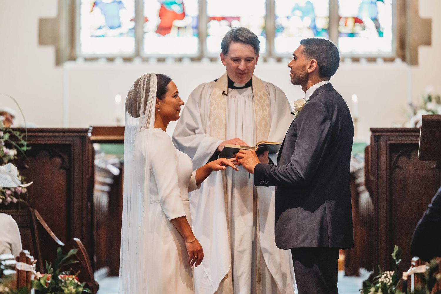 A couple exchanging rings during their wedding ceremony in a church with stained glass windows and floral decorations.
