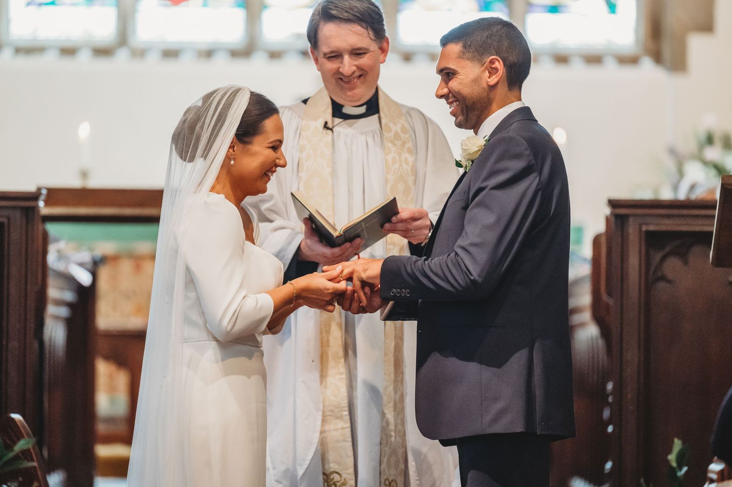 A wedding ceremony takes place in a church as a couple exchanges rings with a robed officiant presiding.