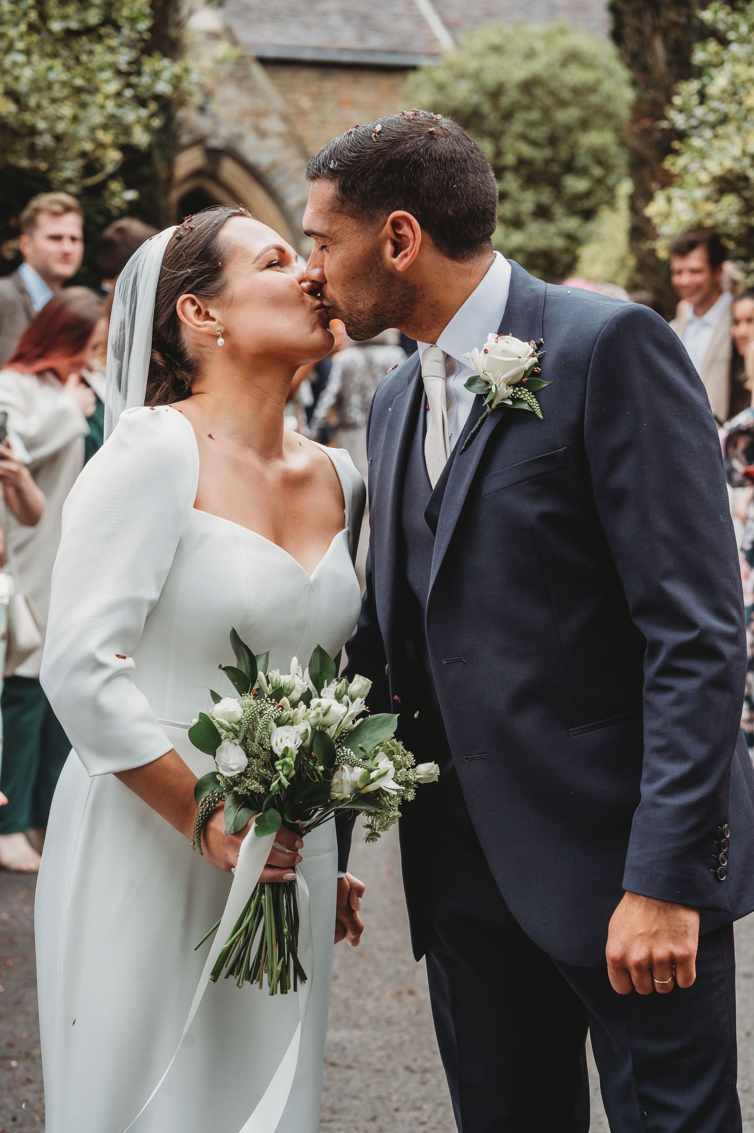 A romantic wedding kiss between the bride in a modern long-sleeved dress and groom in navy suit holding a white bouquet.