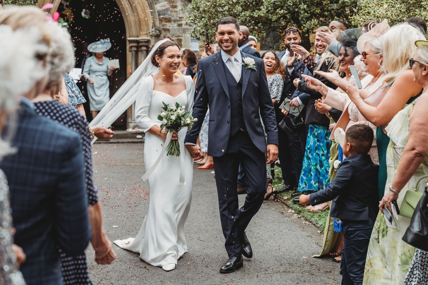 Wedding couple exits church ceremony while guests throw confetti and take photos along pathway.