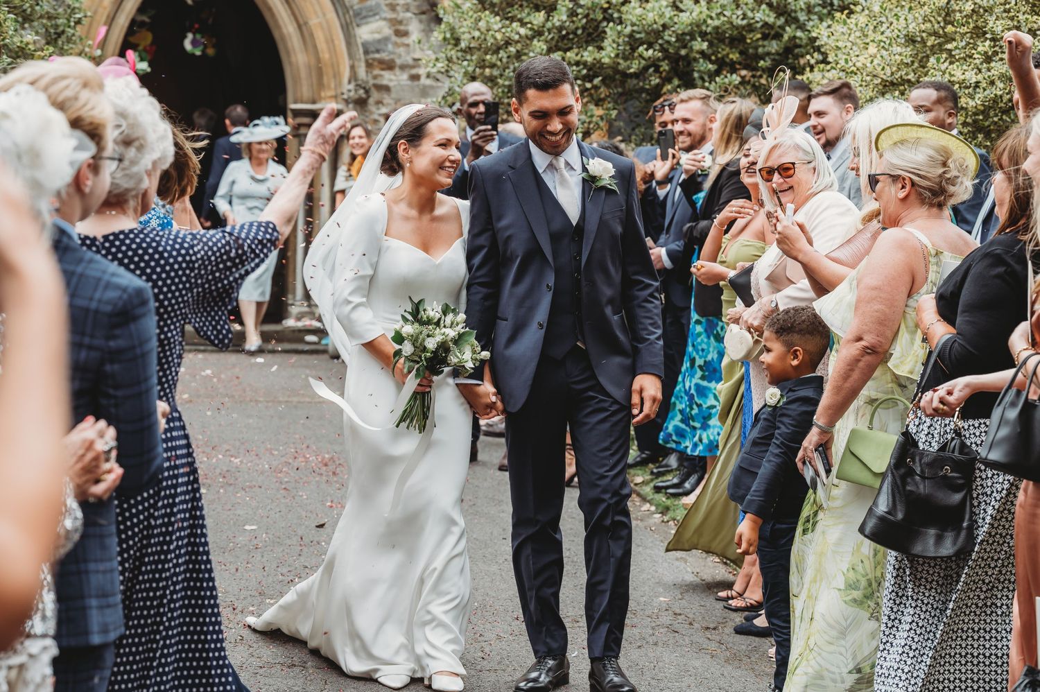 Wedding couple walks down path between rows of celebratory guests tossing confetti after outdoor ceremony.