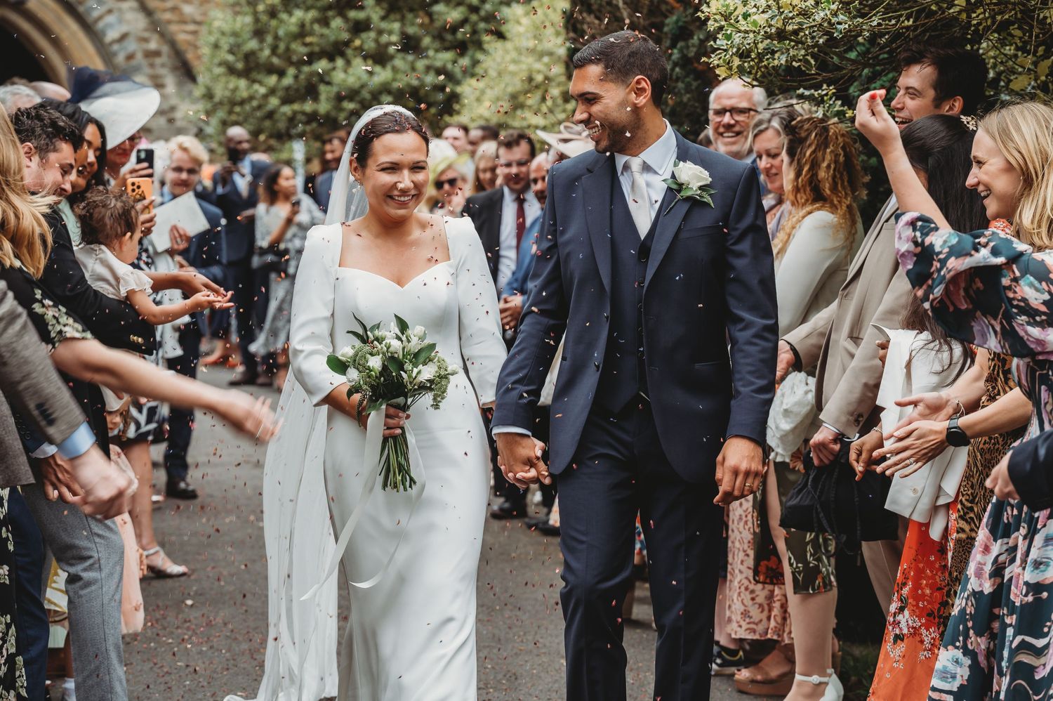 Wedding couple walks through crowd of celebrating guests throwing confetti along pathway at outdoor ceremony.