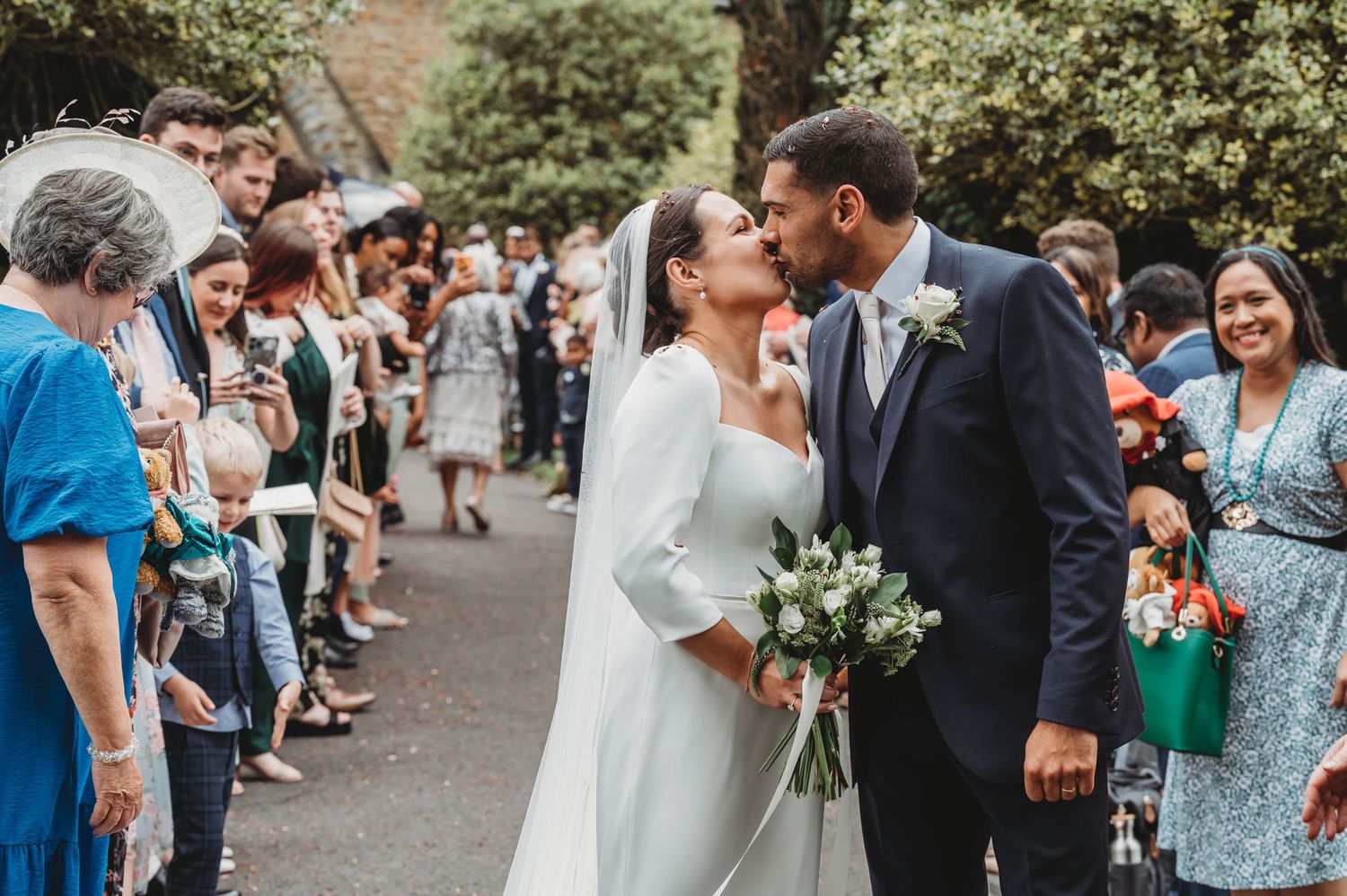 Two newlyweds share an intimate moment surrounded by guests at an outdoor wedding ceremony.