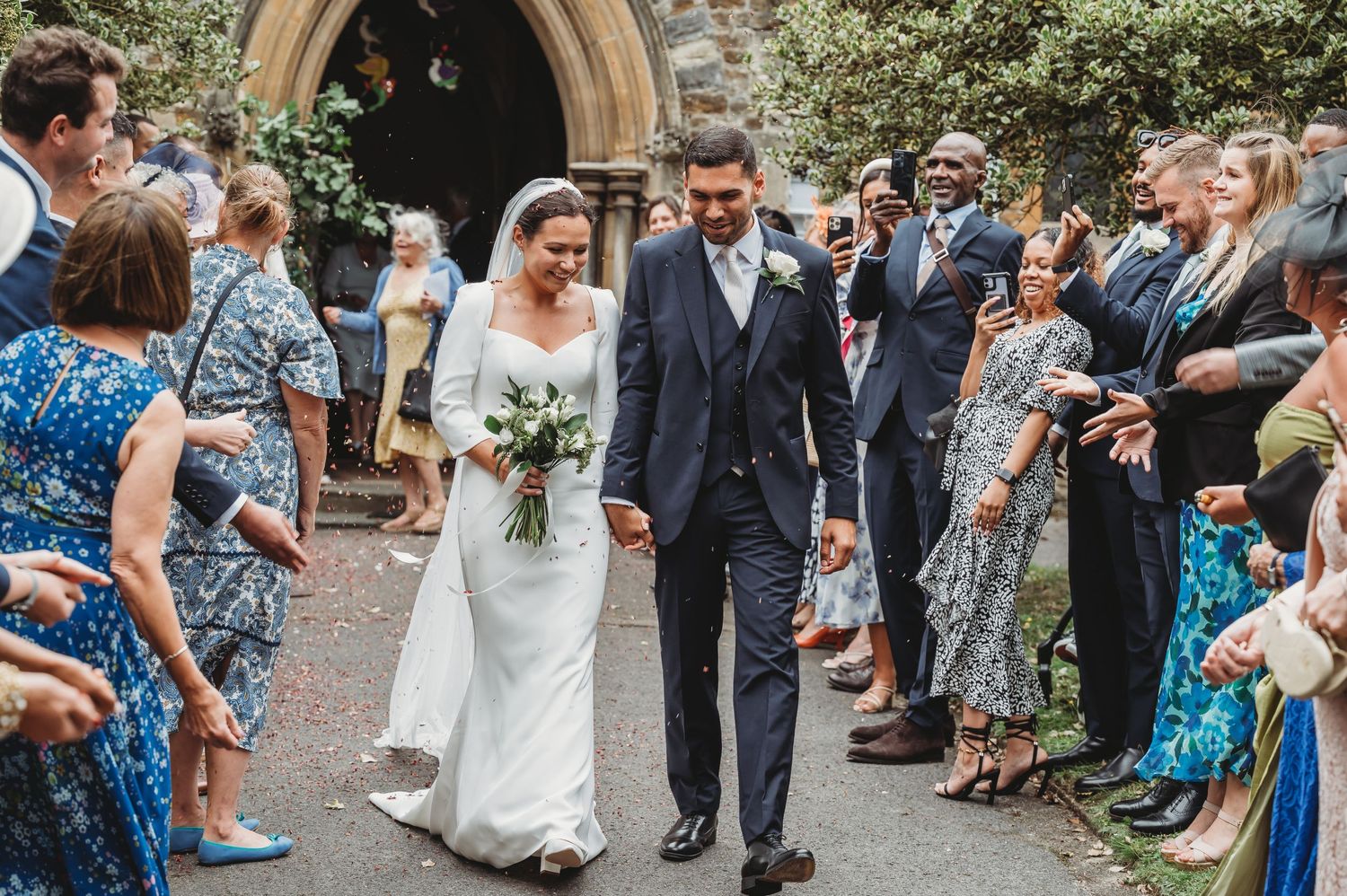 Wedding guests line a church path with confetti as a couple in white dress and navy suit exit the religious ceremony.