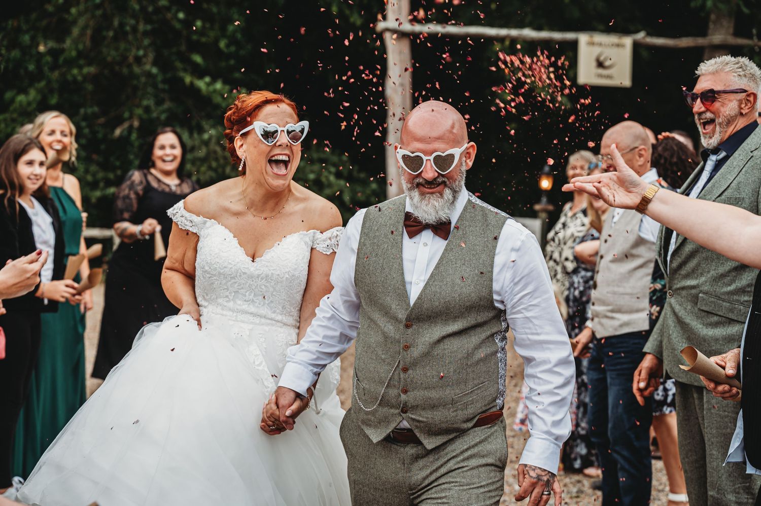 A joyful couple in wedding attire smiles as guests throw confetti during an outdoor wedding celebration.