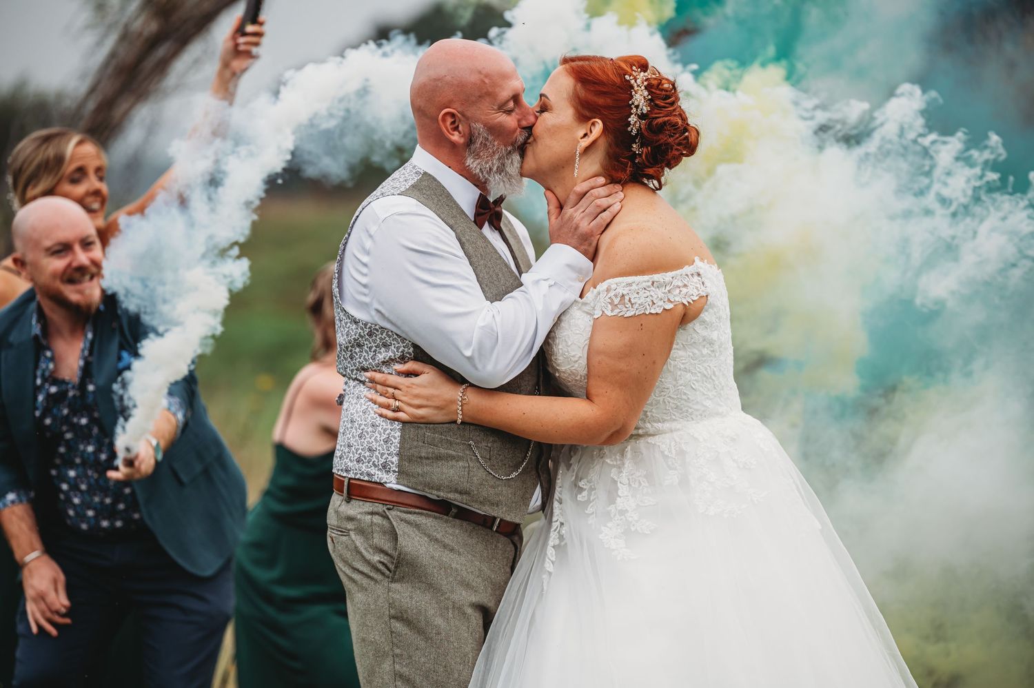 A couple shares a romantic kiss surrounded by turquoise smoke bombs at their outdoor wedding celebration.
