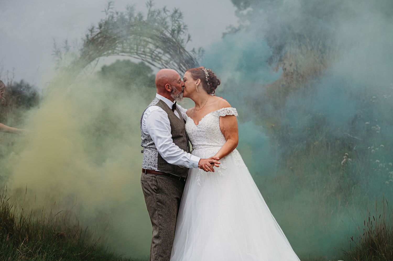 A romantic wedding moment captured with vibrant smoke bombs creating teal and yellow clouds in the background.