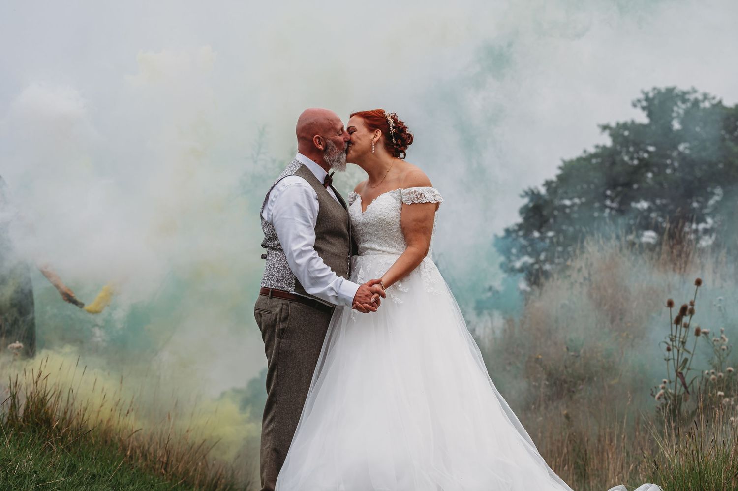 Newlyweds embrace in a dreamy outdoor setting with colorful smoke effects and tall grass in the background.