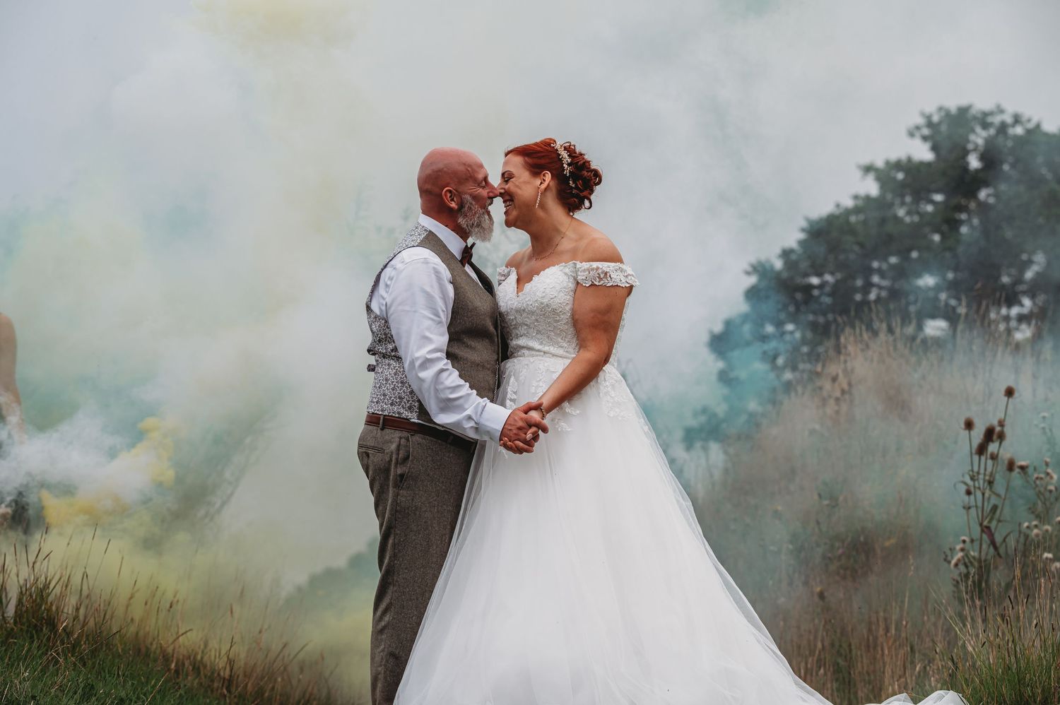 A wedding couple embraces against a backdrop of colorful smoke in a dreamy outdoor setting.