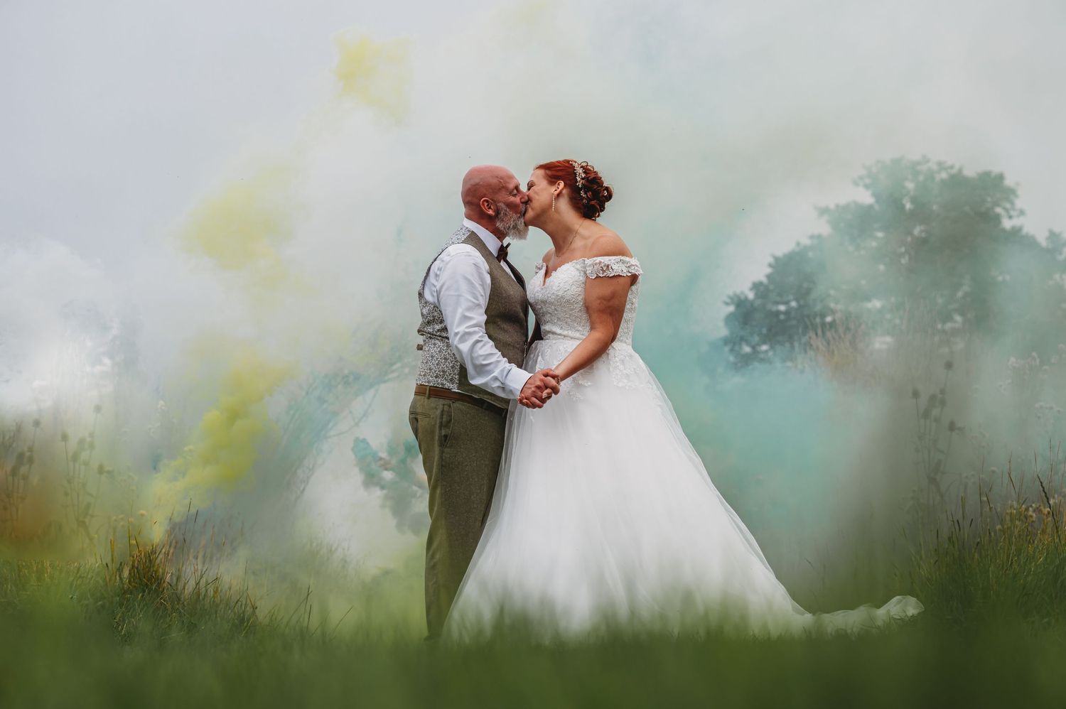 A couple shares a romantic moment in a misty field wearing wedding attire with soft colorful smoke effects in the background.