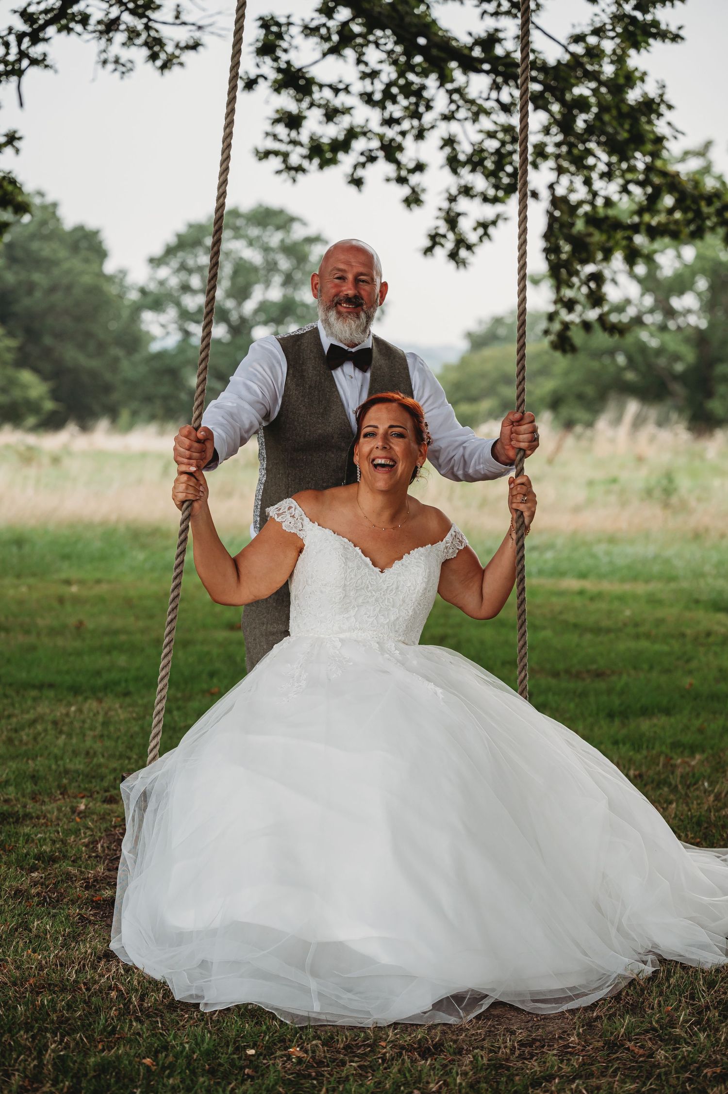 A joyful wedding couple shares a sweet moment together on a rustic rope swing outdoors under tree branches.