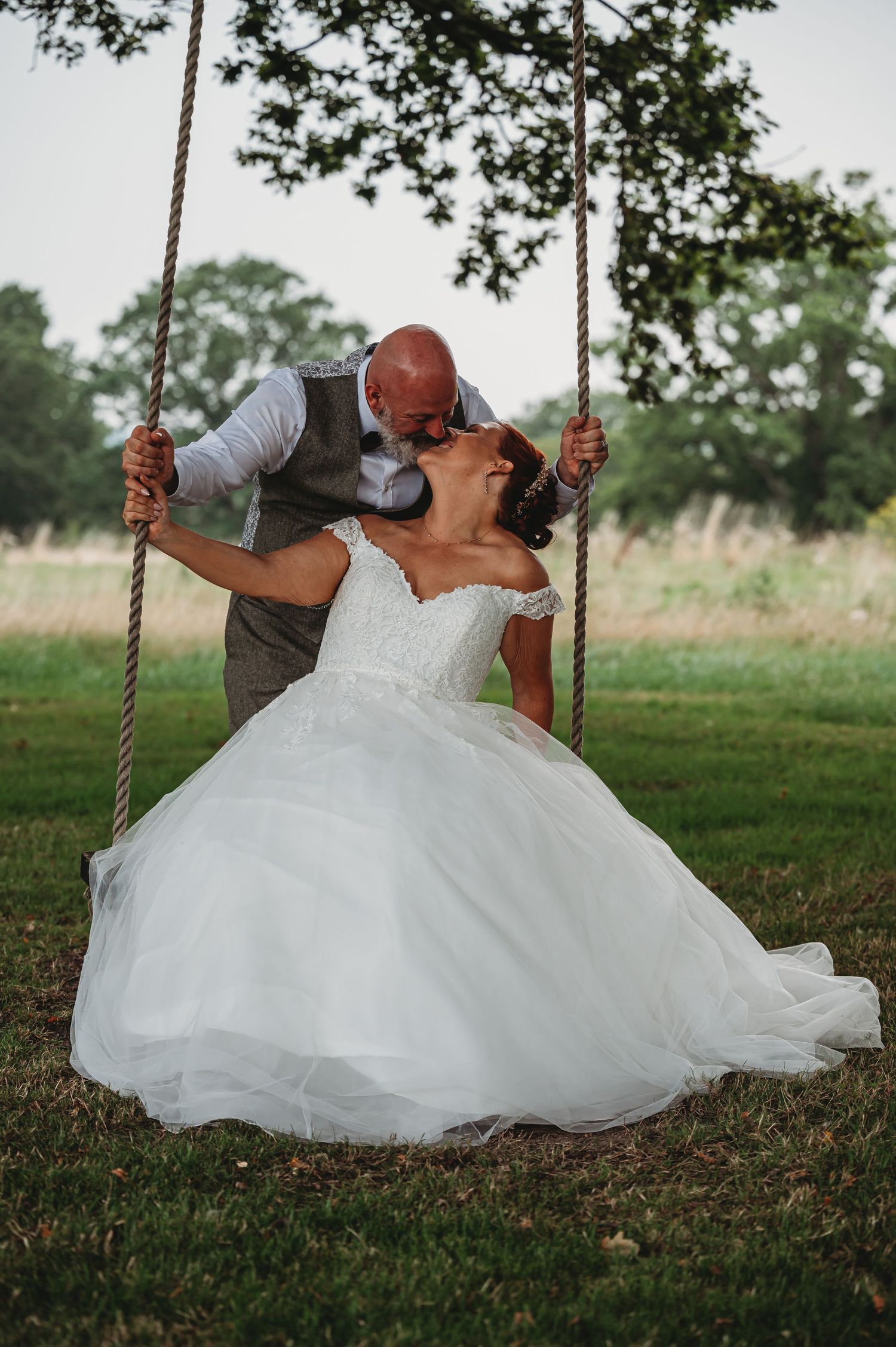 A romantic wedding moment captured outdoors with a couple sharing a kiss while the bride sits on a rustic rope swing.