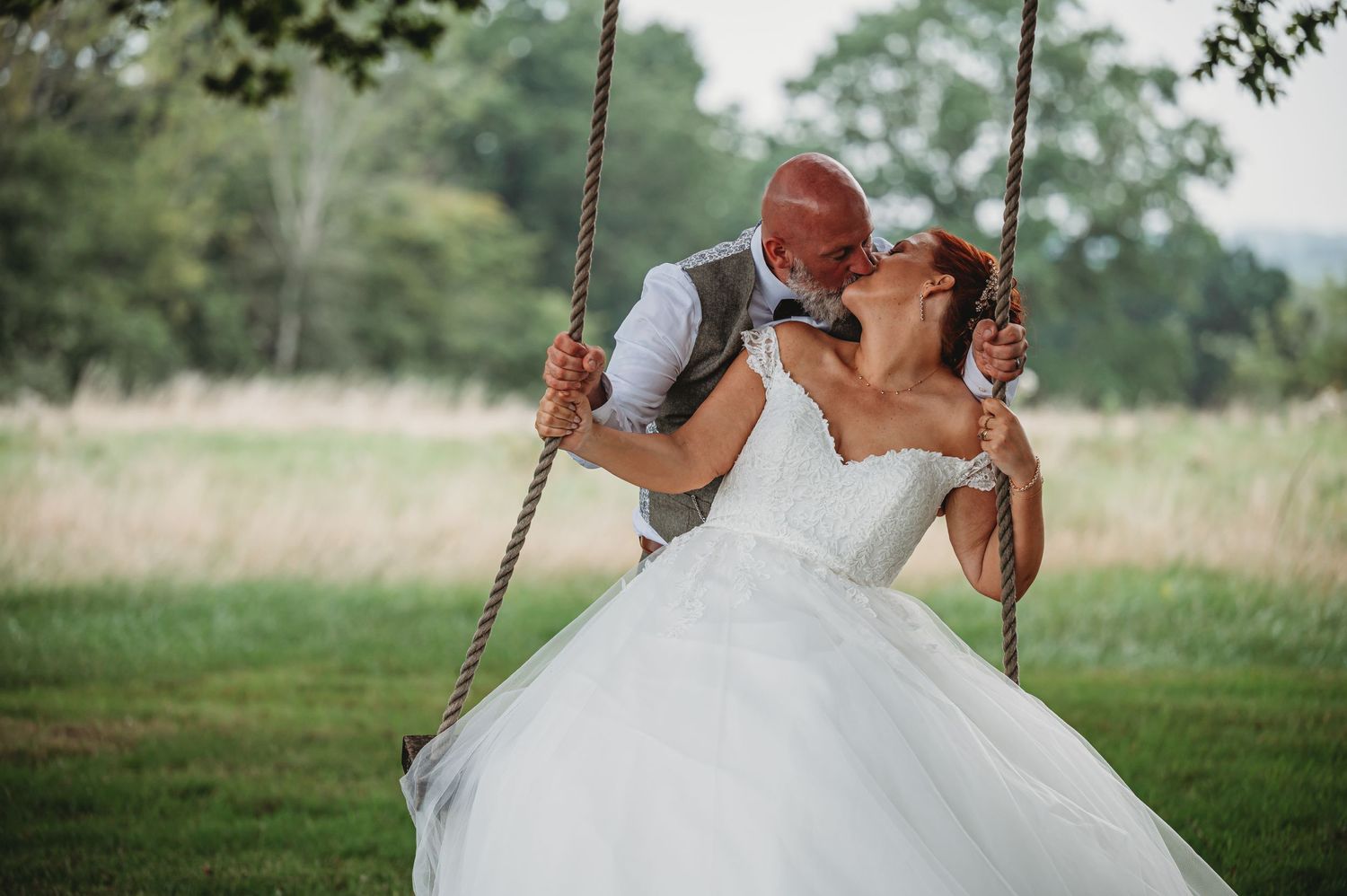 A romantic couple shares a sweet kiss while sitting on a rustic rope swing in a beautiful outdoor country setting.