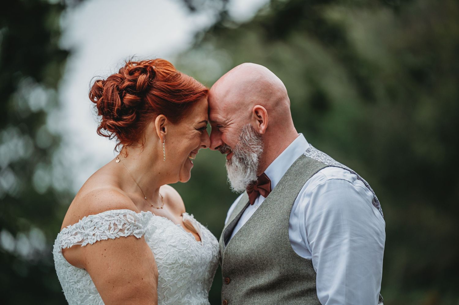 Couple shares an intimate moment outdoors during their wedding celebration, with bride in lace dress and groom in gray vest.