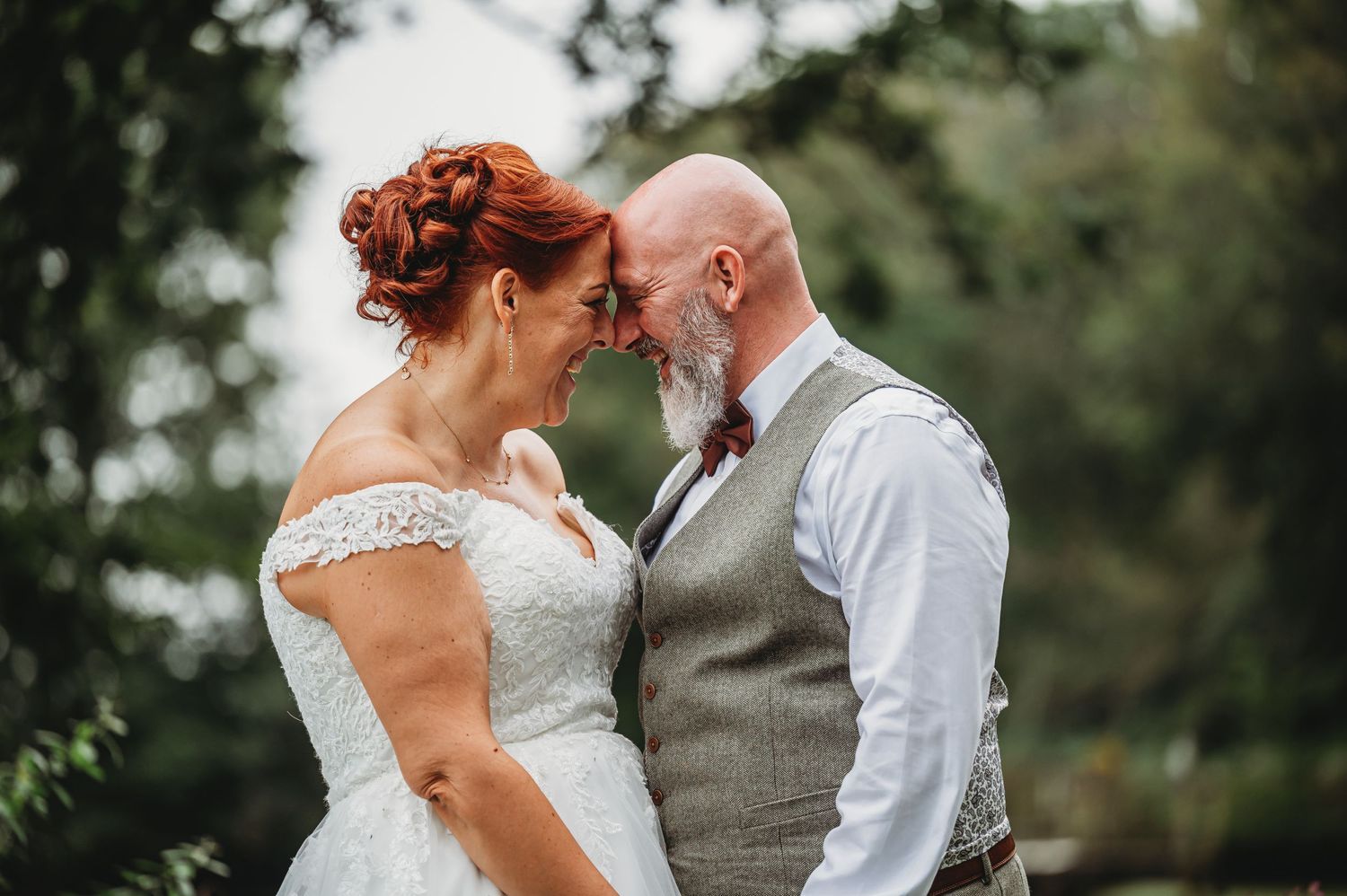 A couple shares an intimate moment outdoors during their wedding, with the bride in lace and groom in a gray vest.