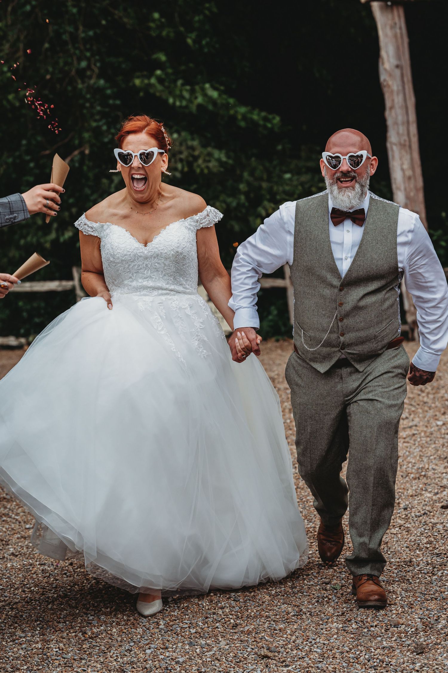 A joyful couple in wedding attire laugh while walking hand in hand on a gravel path surrounded by trees.