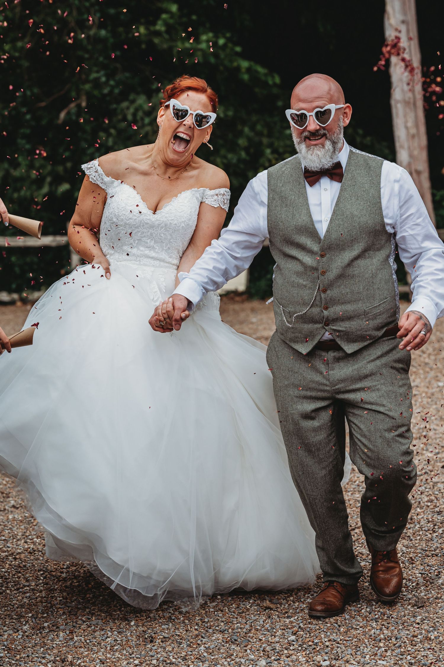 A joyful wedding couple wearing sunglasses share a candid moment on a gravel path, with the bride in a flowing white dress.