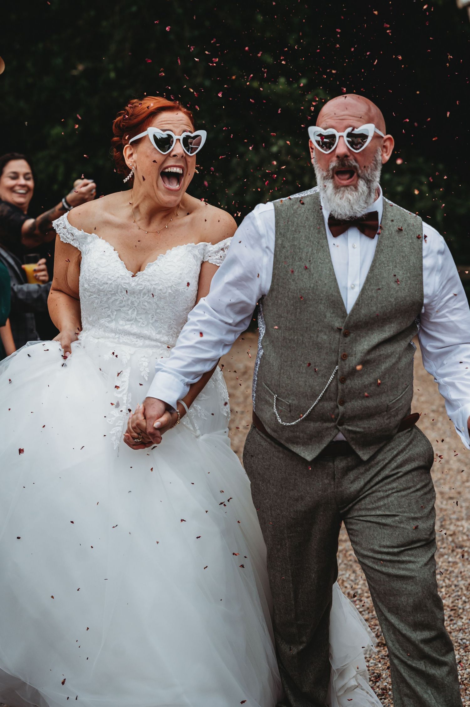 Happy newlyweds in matching white sunglasses laugh and hold hands while walking through falling confetti.