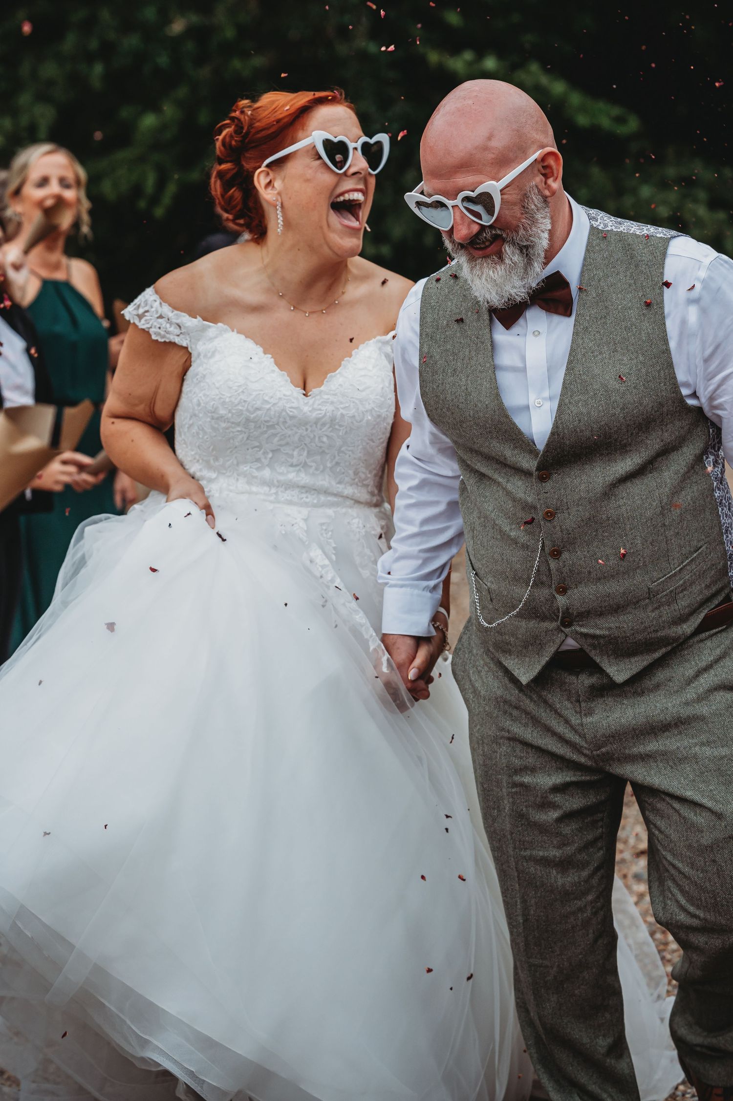 A joyful wedding couple wearing matching sunglasses walk together as confetti falls around them.
