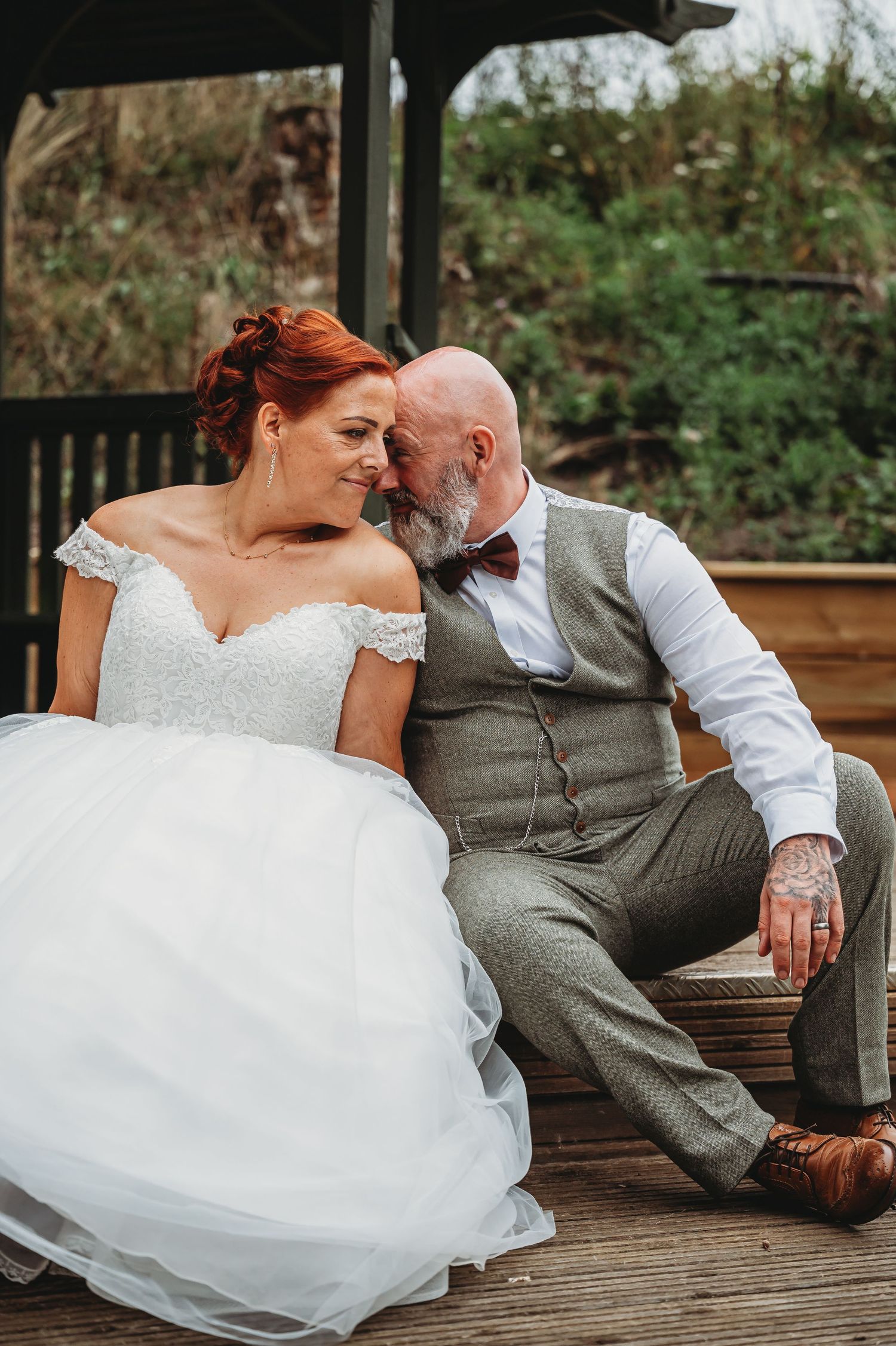 A lovely wedding couple sitting on a wooden deck, with the bride in white and the groom in a gray vest and bowtie.