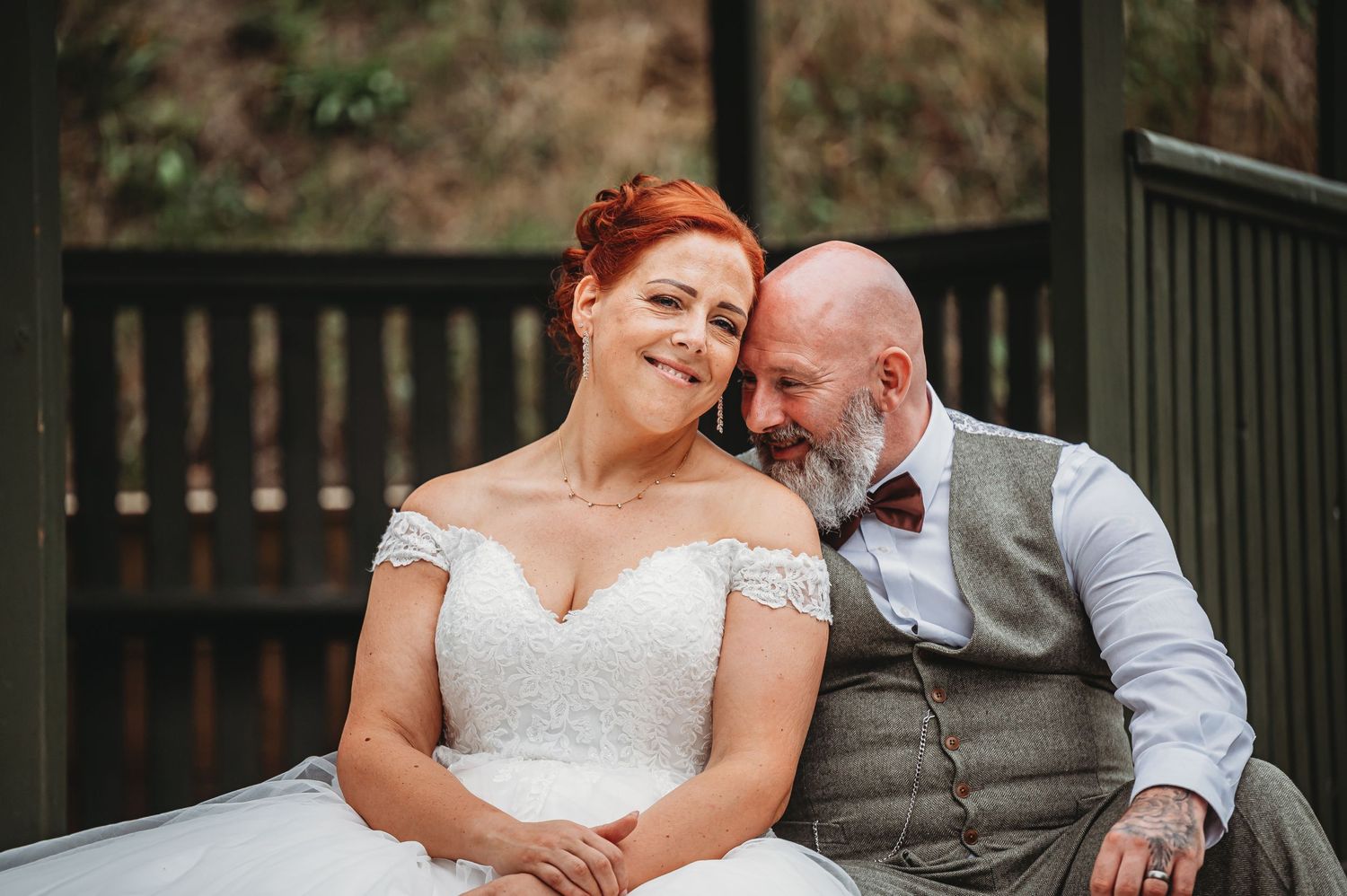 A couple in wedding attire shares a tender moment while sitting together outdoors on their special day.