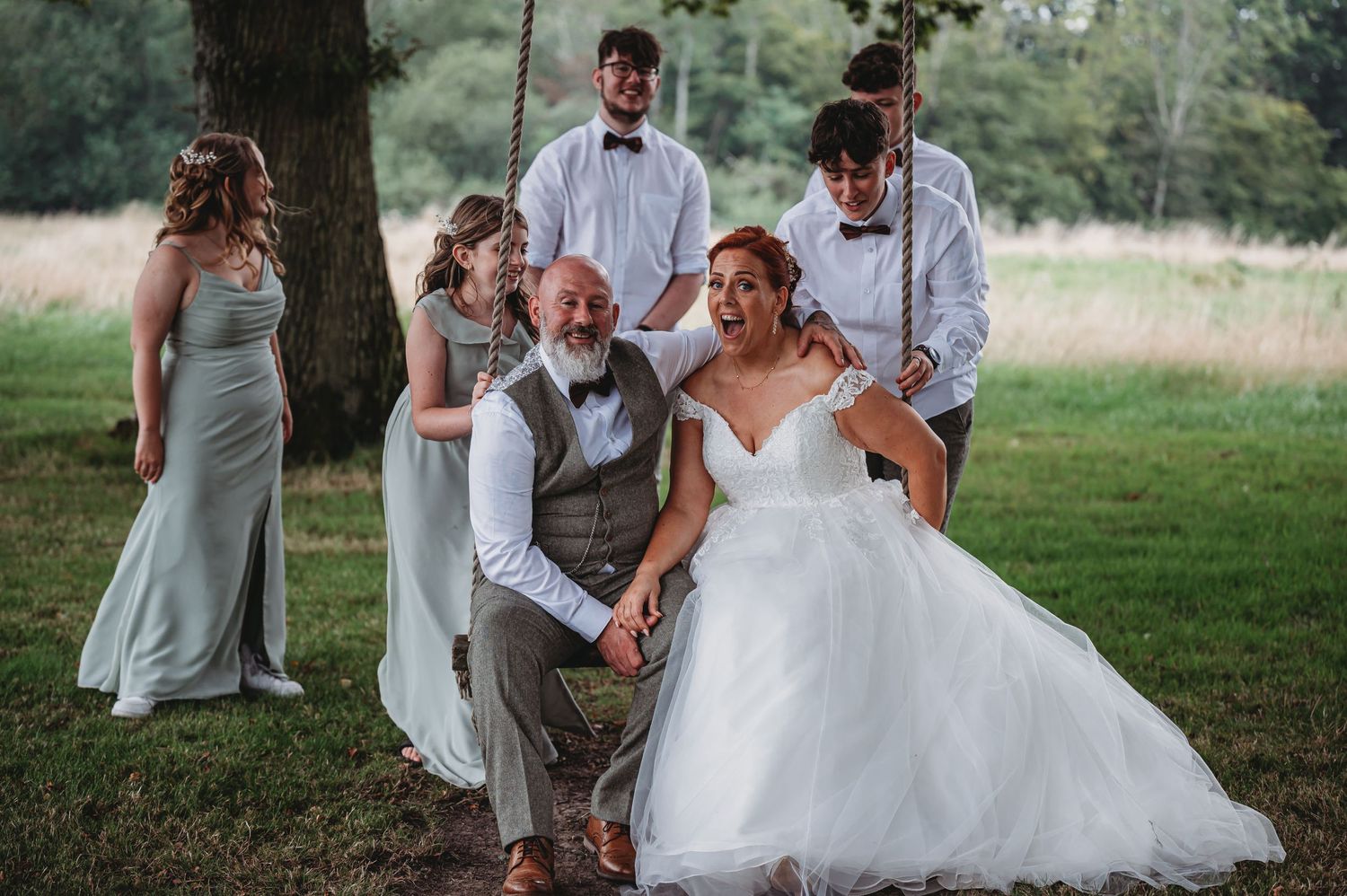 A bridal party shares a candid moment together on a grassy lawn under a tree during an outdoor wedding celebration.
