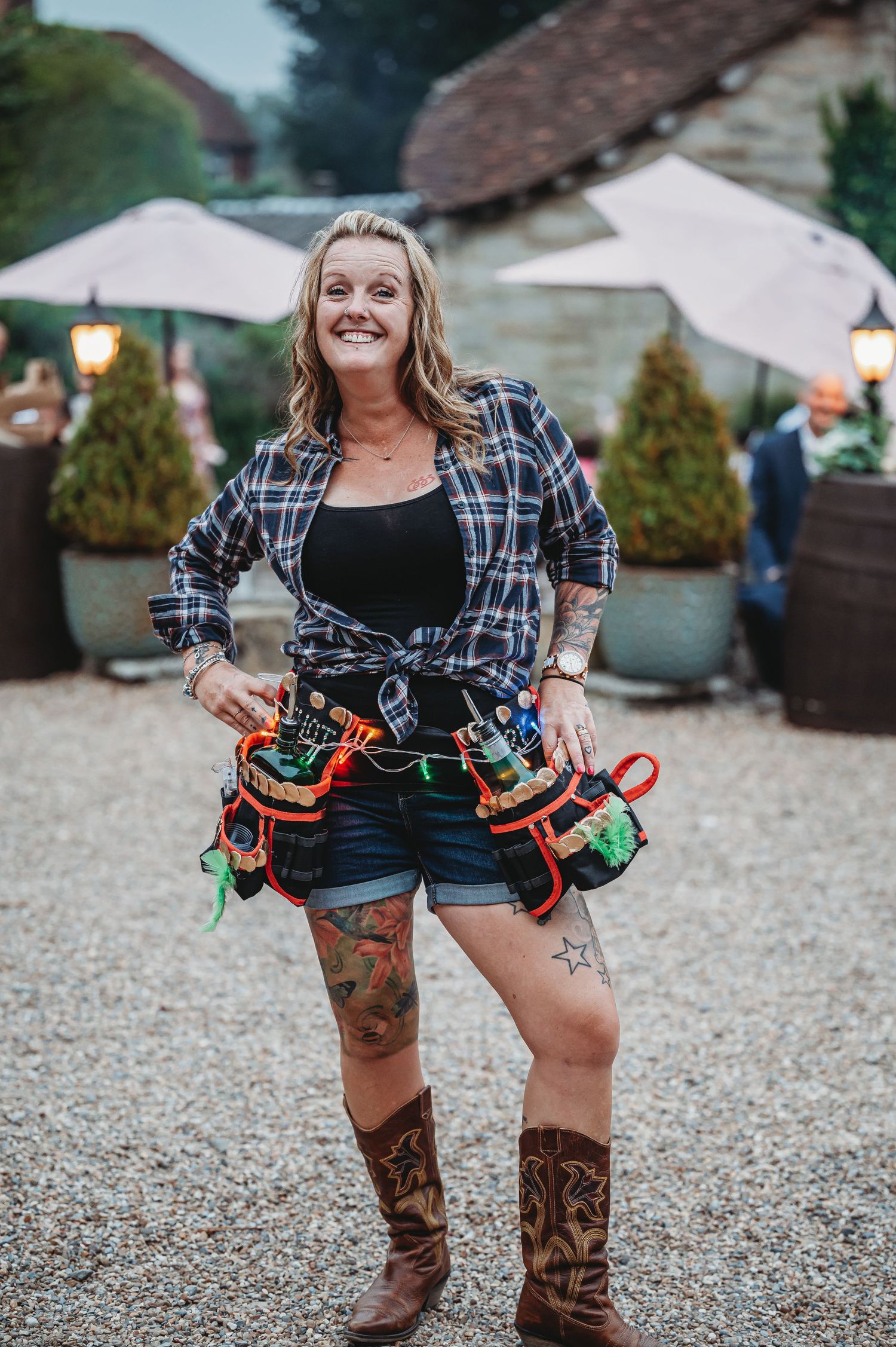 Worker wearing plaid shirt and cowboy boots with tool belt stands on gravel at outdoor event with string lights visible.