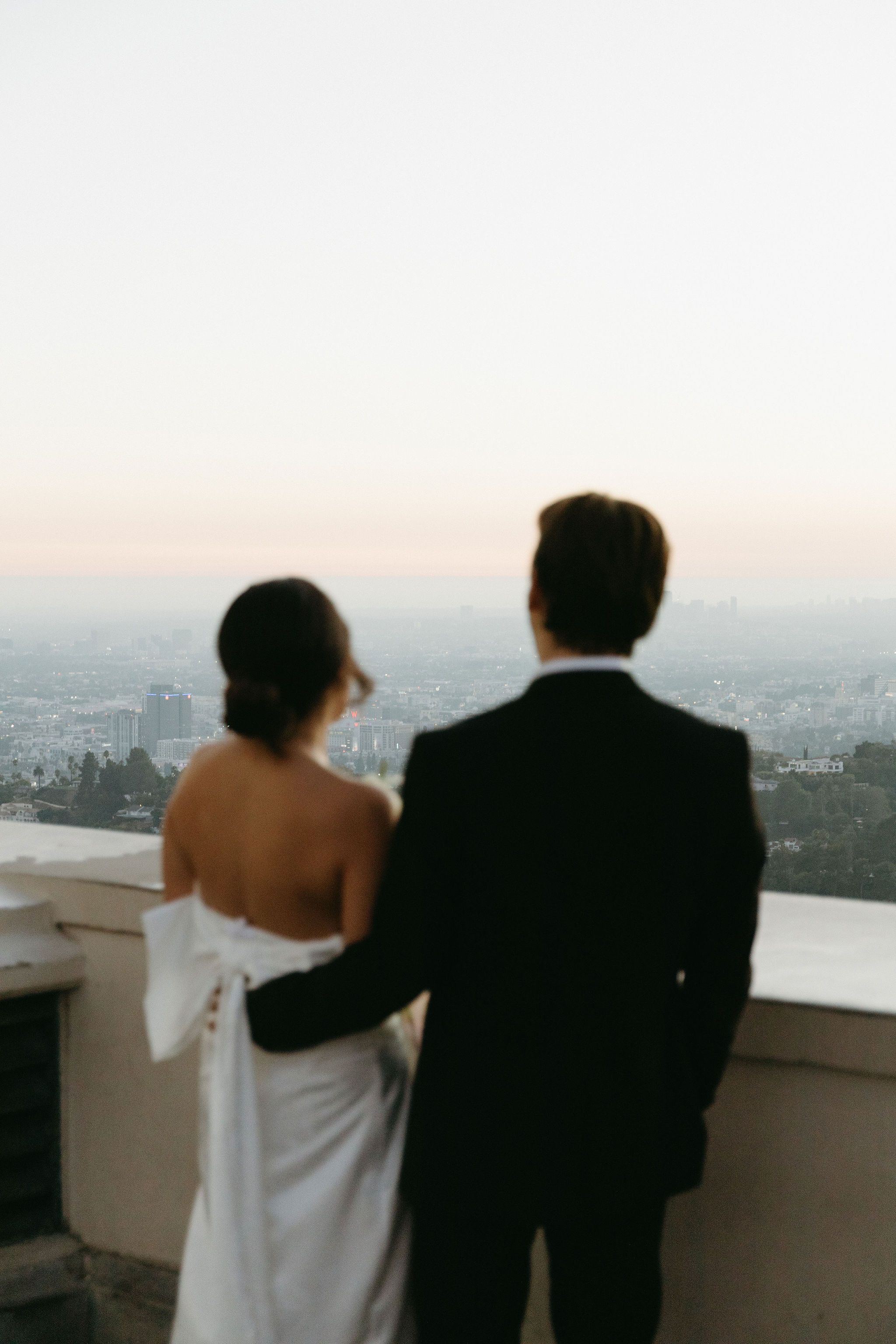 Couple in formal attire stand on balcony overlooking city skyline at sunset.