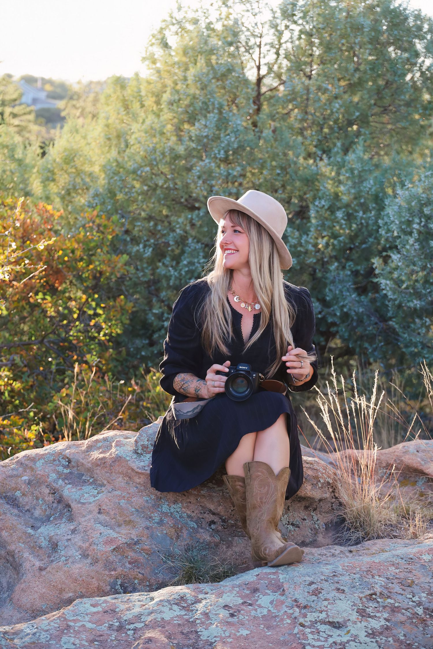Person in black dress and hat sitting on red rocks with camera during golden hour.