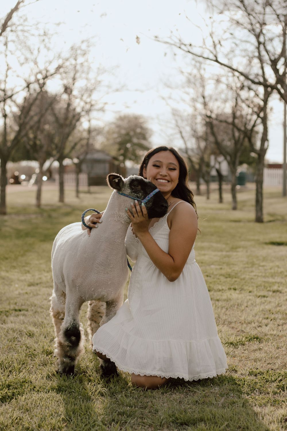 A person in a white dress plays with a small white dog on grassy lawn during a photoshoot.