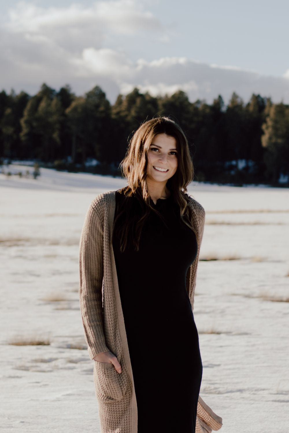 A winter portrait of someone wearing a long cardigan and black dress in snowy landscape.