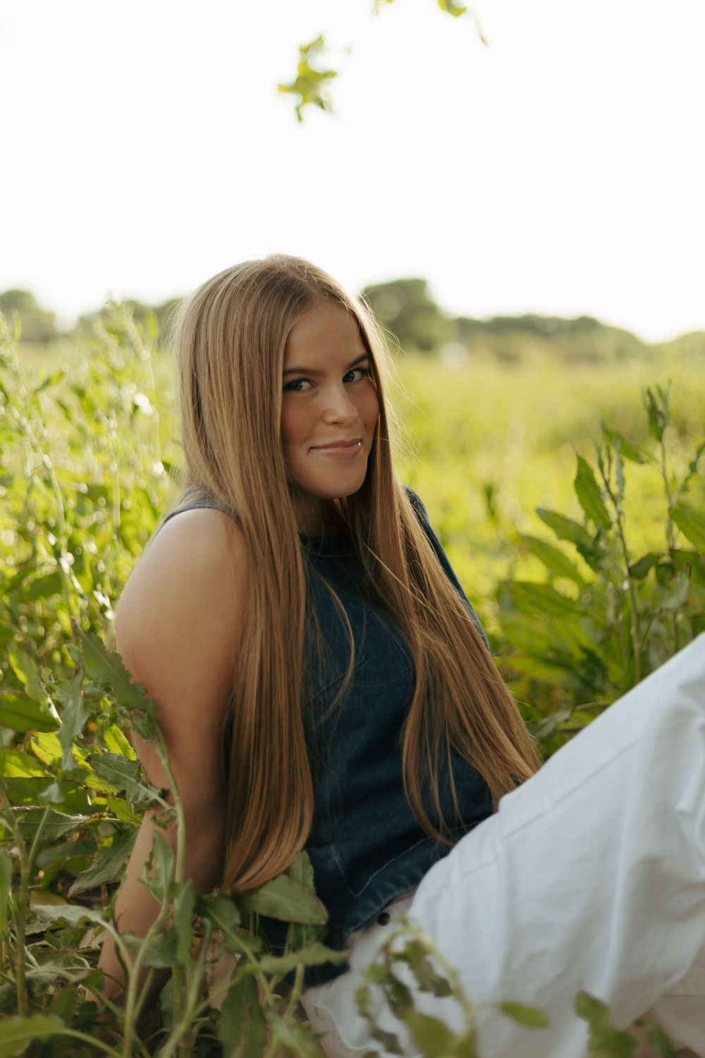 A woman with long hair sits in a green field wearing a navy blue top and white pants.