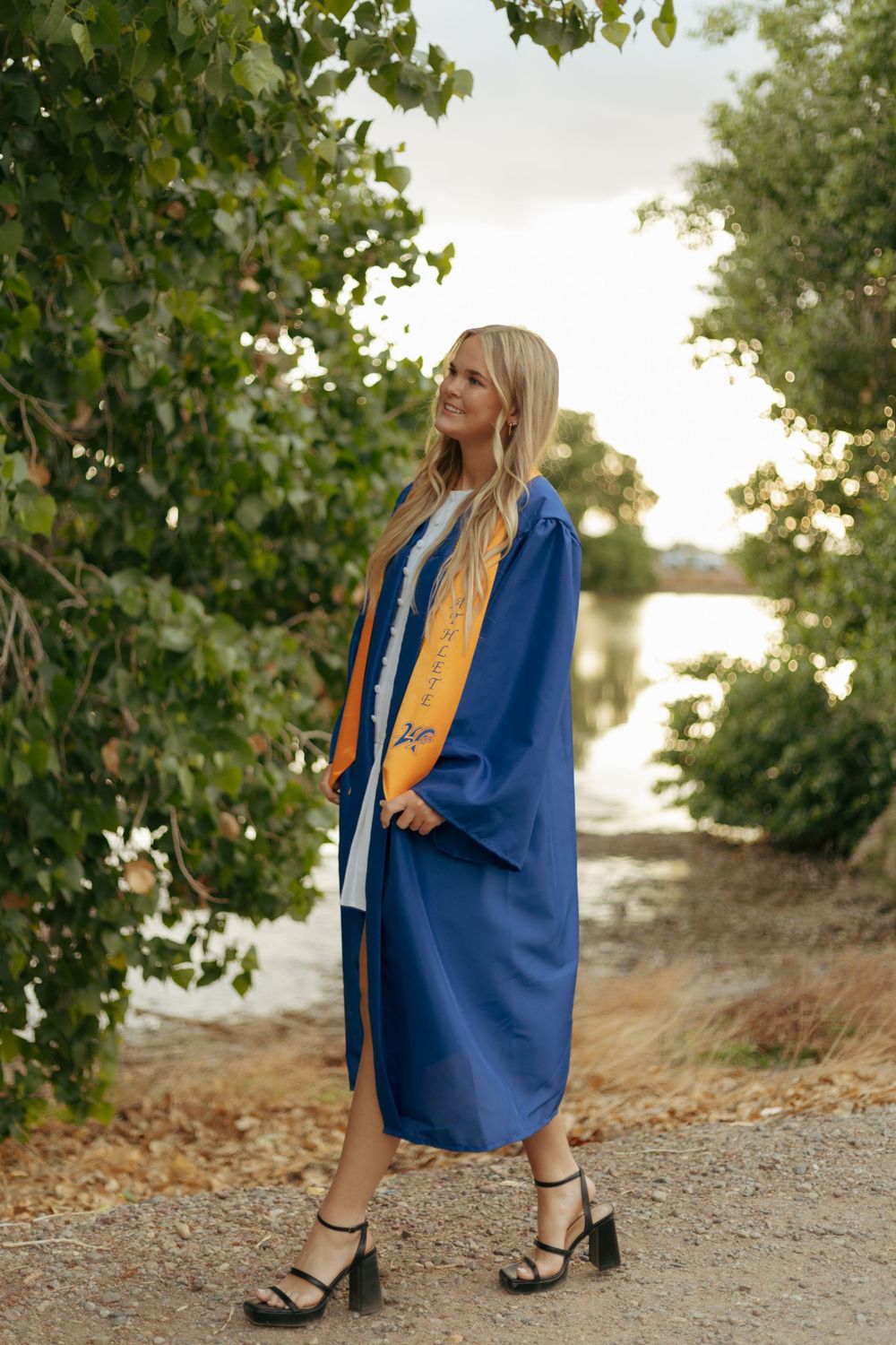 A graduate in blue and gold regalia poses on a path near water during golden hour.