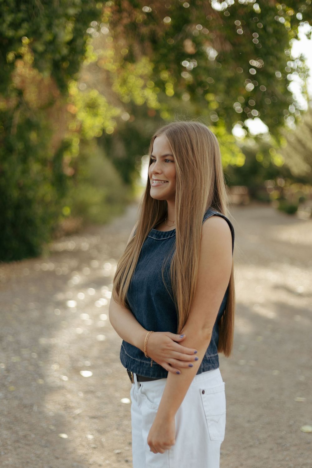 A woman in a navy sleeveless top and white pants poses on a sunlit dirt path surrounded by greenery.