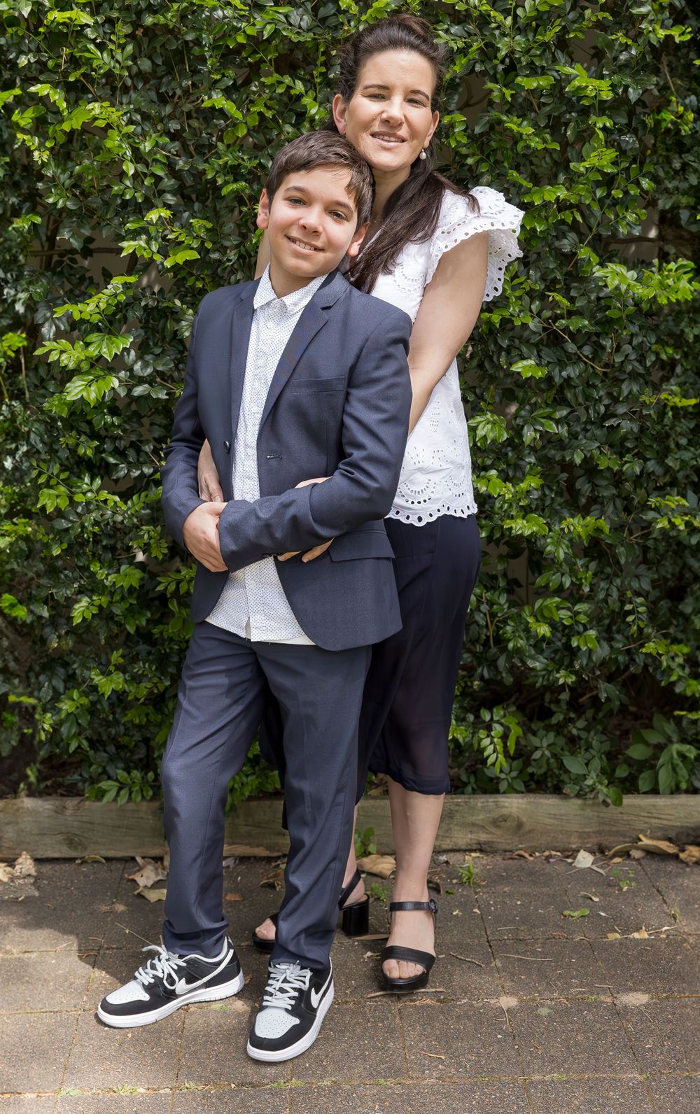 Formal portrait of two people standing together against green hedge background.