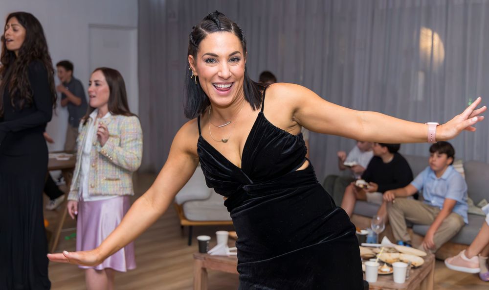 Person in a black velvet dress dancing with arms outstretched at an indoor event.