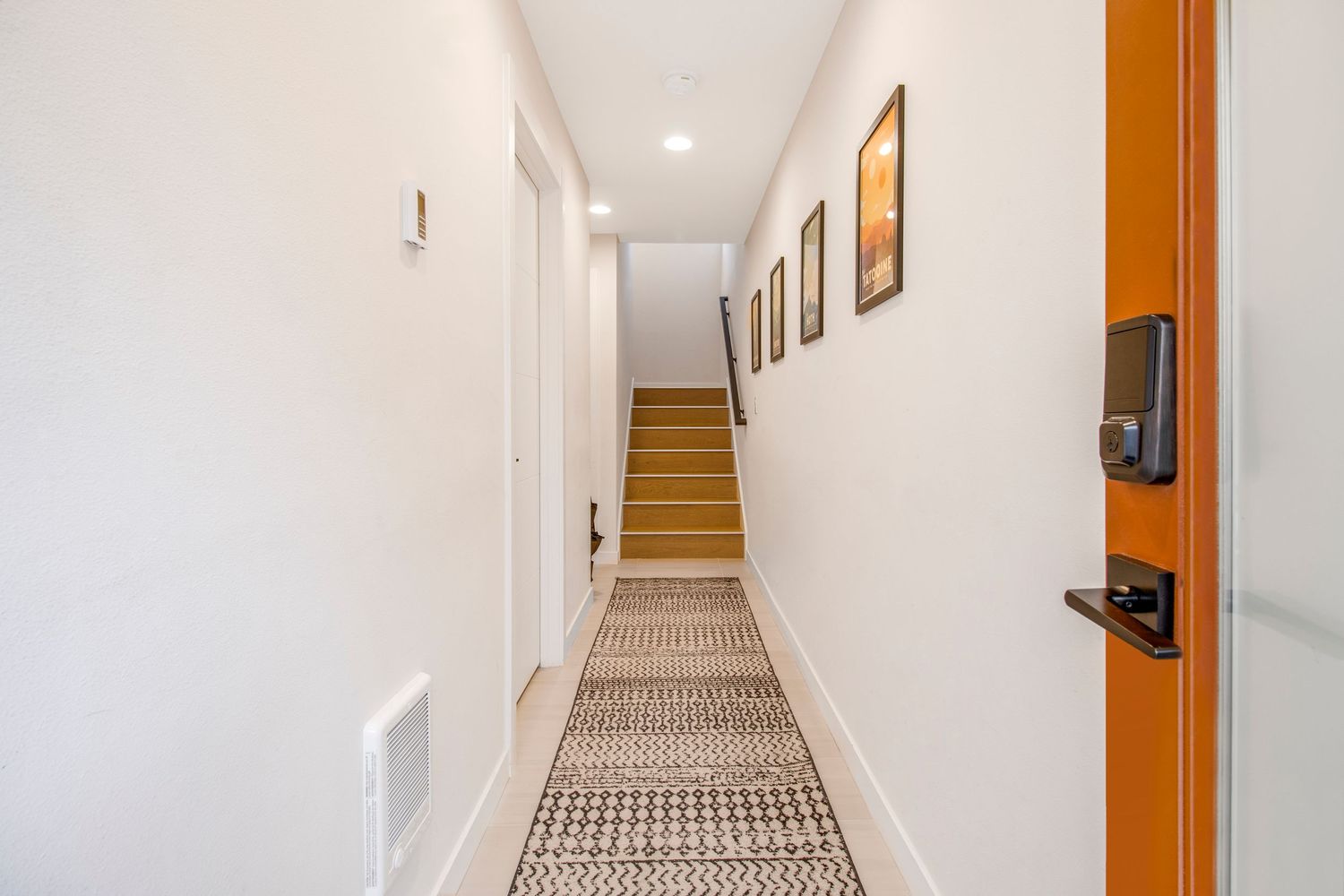 Long hallway with patterned carpet runner, orange door, and framed artwork on white walls.